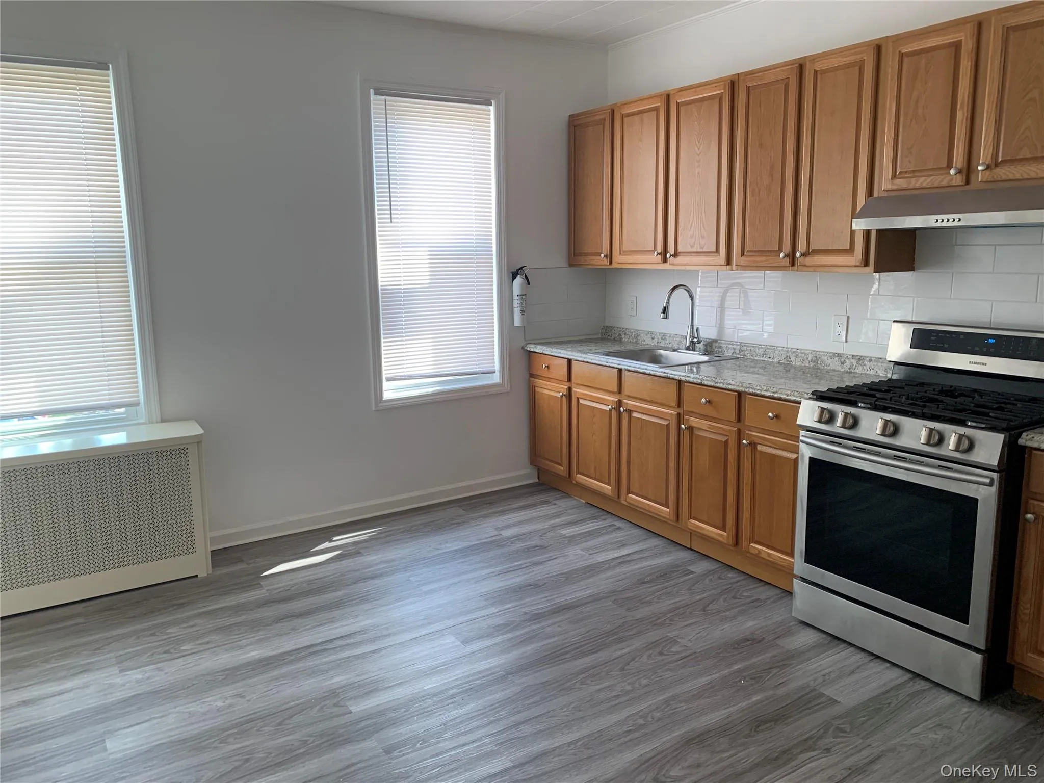 Kitchen featuring stainless steel gas stove, radiator, backsplash, brown cabinetry, and dark wood-style flooring Kitchen featuring stainless steel gas stove, radiator, backsplash, brown cabinetry, and dark wood-style flooring