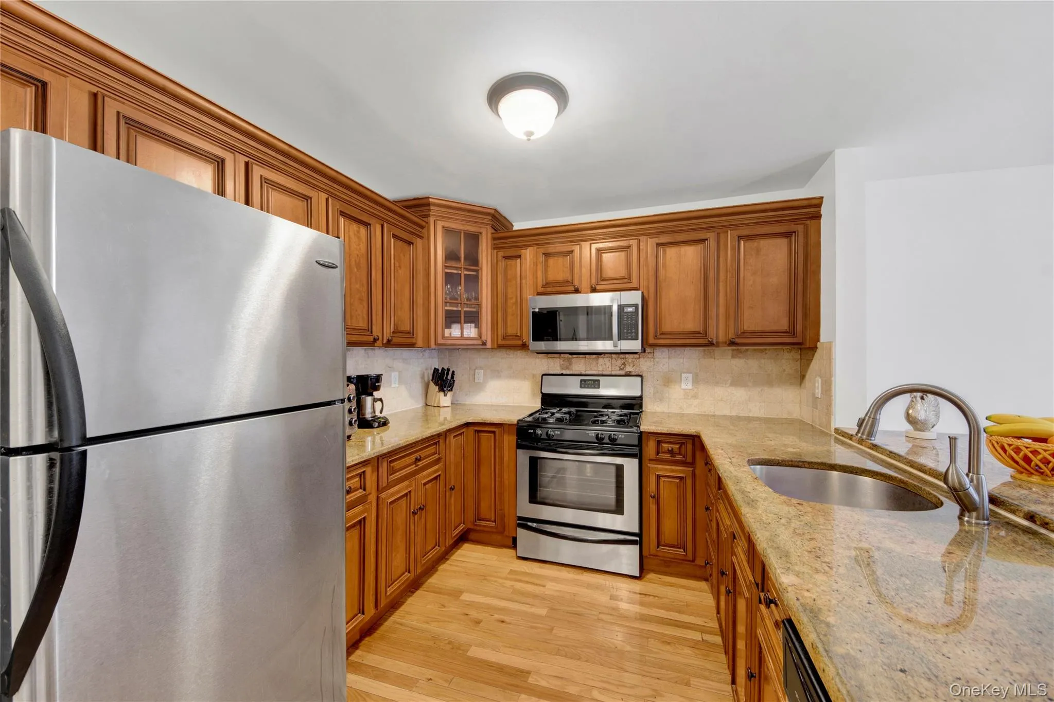 Kitchen with appliances with stainless steel finishes, brown cabinets, light stone counters, light wood finished floors, and decorative backsplash Kitchen with appliances with stainless steel finishes, brown cabinets, light stone counters, light wood finished floors, and decorative backsplash