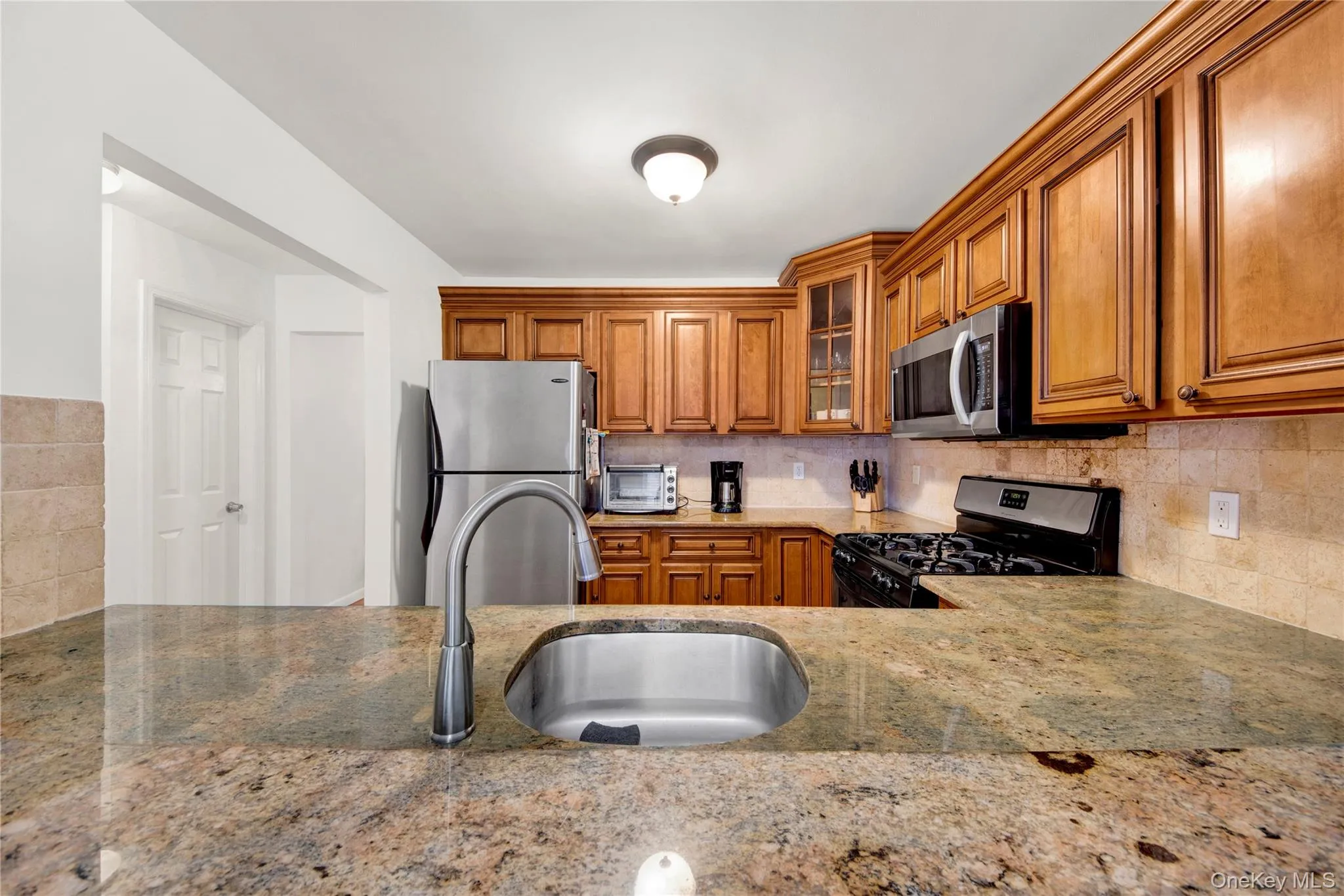 Kitchen with brown cabinets, glass insert cabinets, appliances with stainless steel finishes, decorative backsplash, and light stone countertops Kitchen with brown cabinets, glass insert cabinets, appliances with stainless steel finishes, decorative backsplash, and light stone countertops