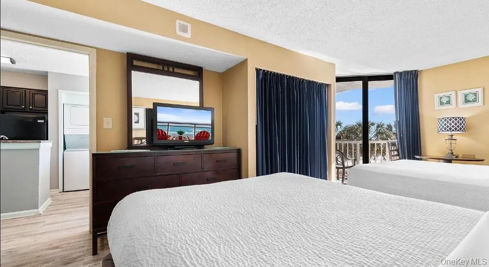 Bedroom featuring a textured ceiling, black fridge, stacked washer / dryer, light wood-type flooring, and access to exterior Bedroom featuring a textured ceiling, black fridge, stacked washer / dryer, light wood-type flooring, and access to exterior