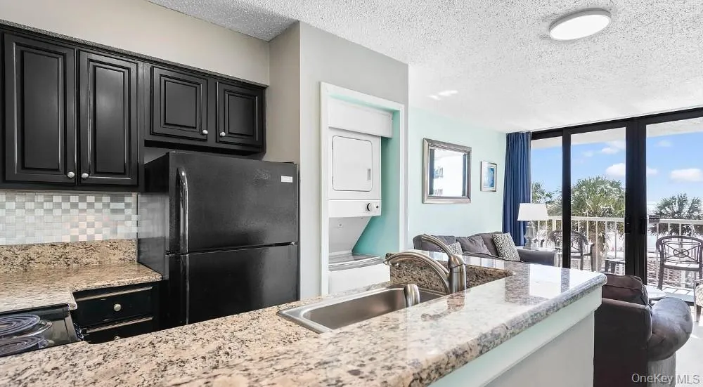 Kitchen with a wall of windows, black appliances, stacked washer and clothes dryer, a textured ceiling, and open floor plan Kitchen with a wall of windows, black appliances, stacked washer and clothes dryer, a textured ceiling, and open floor plan