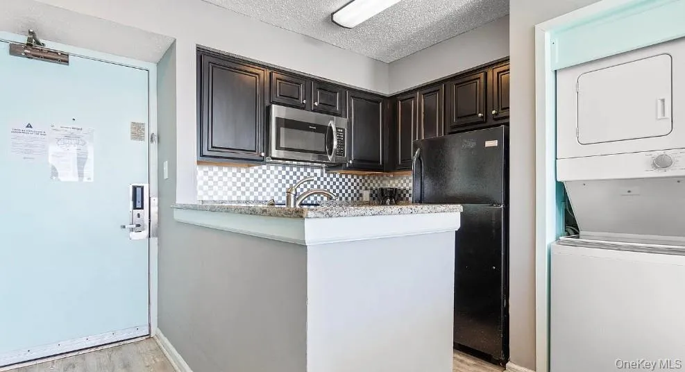 Kitchen featuring freestanding refrigerator, a textured ceiling, tasteful backsplash, stacked washer / dryer, and stainless steel microwave Kitchen featuring freestanding refrigerator, a textured ceiling, tasteful backsplash, stacked washer / dryer, and stainless steel microwave