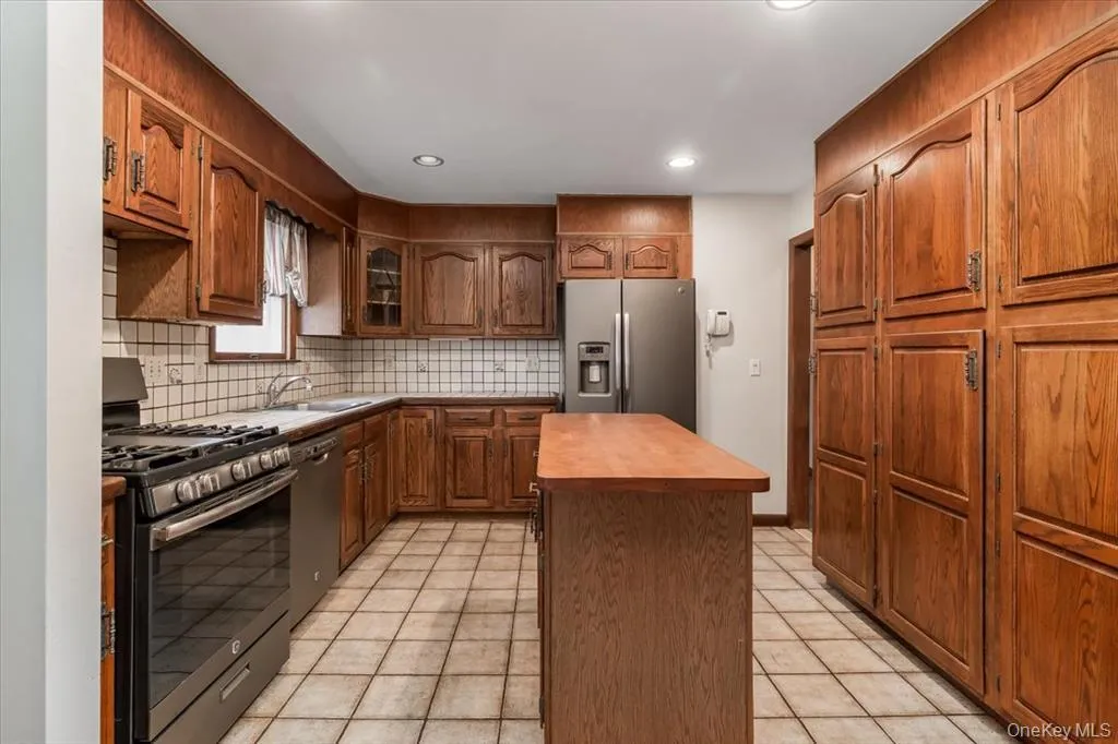Kitchen with stainless steel appliances, brown cabinetry, decorative backsplash, a center island, and light tile patterned floors Kitchen with stainless steel appliances, brown cabinetry, decorative backsplash, a center island, and light tile patterned floors