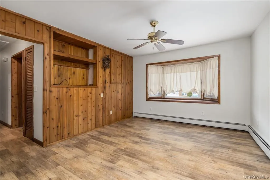 Unfurnished living room featuring wood walls, a baseboard heating unit, light wood-style floors, and ceiling fan Unfurnished living room featuring wood walls, a baseboard heating unit, light wood-style floors, and ceiling fan