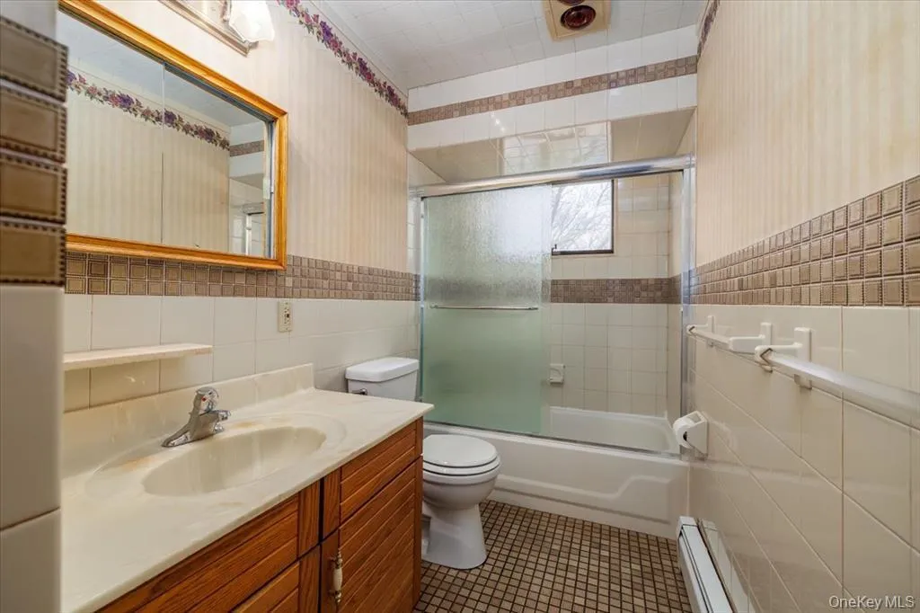 Full bath featuring combined bath / shower with glass door, wallpapered walls, tile walls, vanity, and a baseboard heating unit Full bath featuring combined bath / shower with glass door, wallpapered walls, tile walls, vanity, and a baseboard heating unit