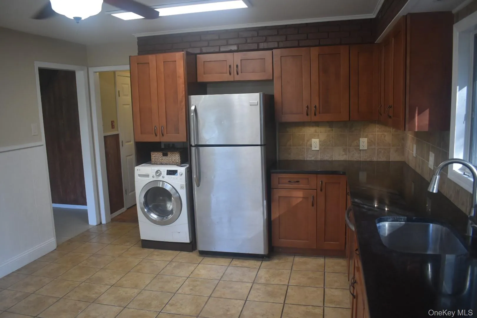 Kitchen featuring freestanding refrigerator, brown cabinetry, washer / clothes dryer, backsplash, and light tile patterned floors Kitchen featuring freestanding refrigerator, brown cabinetry, washer / clothes dryer, backsplash, and light tile patterned floors