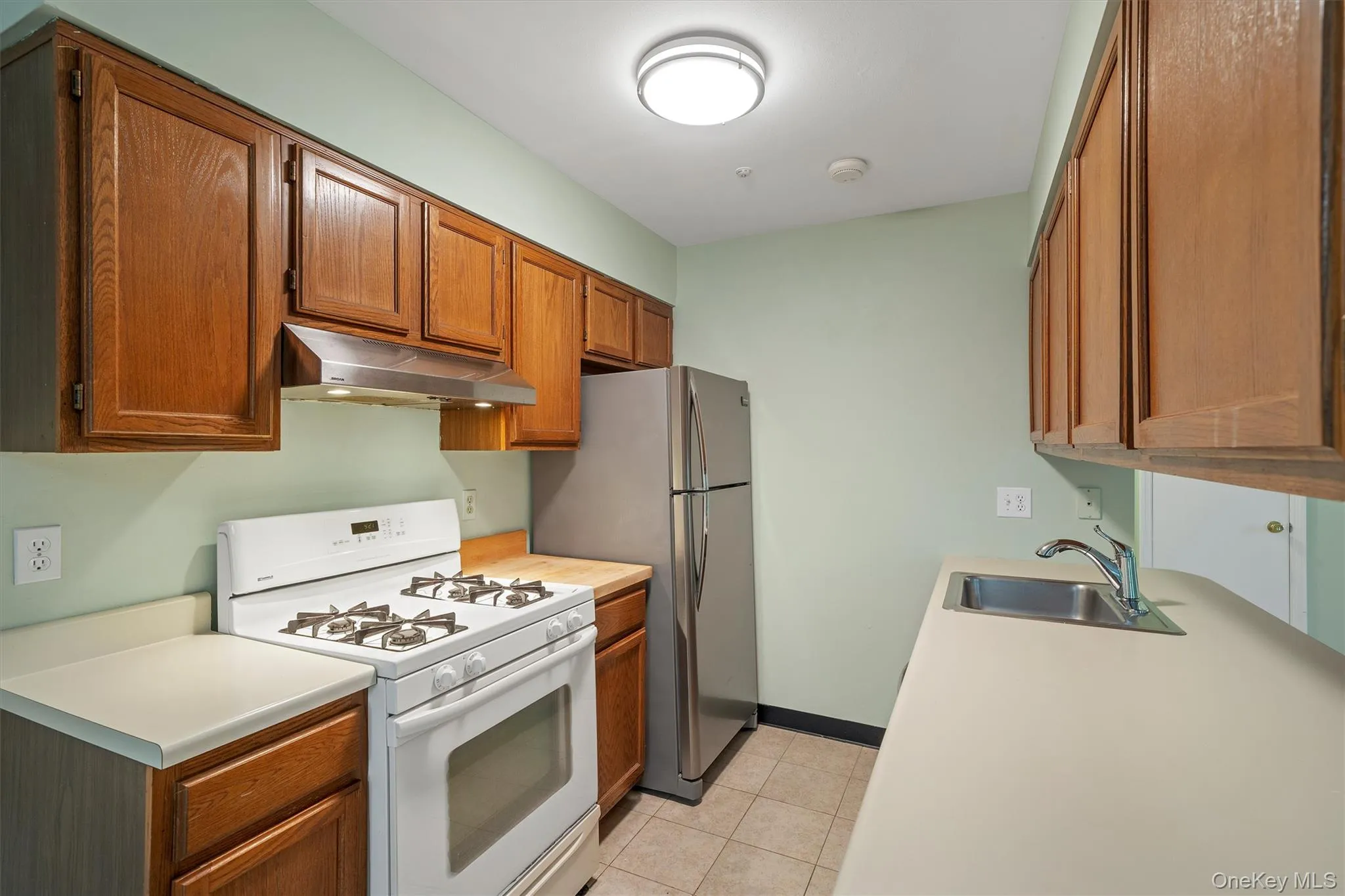 Kitchen with white gas stove, light countertops, under cabinet range hood, and wood cabinetry Kitchen with white gas stove, light countertops, under cabinet range hood, and wood cabinetry