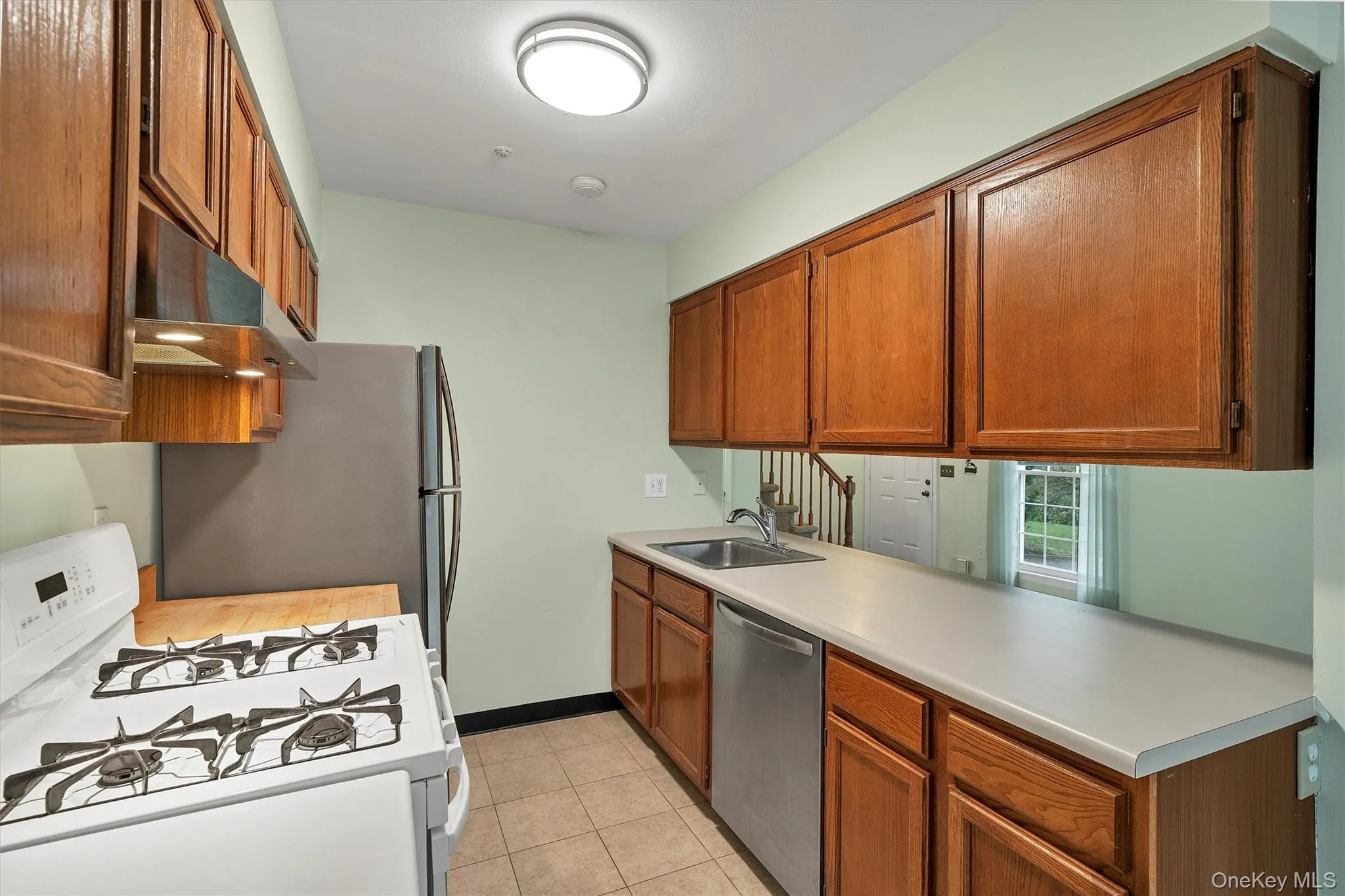 Kitchen with appliances with stainless steel finishes, brown cabinets, light countertops, light tile patterned floors, and under cabinet range hood Kitchen with appliances with stainless steel finishes, brown cabinets, light countertops, light tile patterned floors, and under cabinet range hood