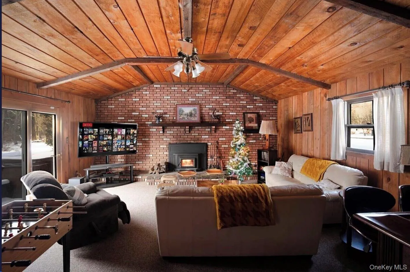Living area featuring wooden walls, wood ceiling, a ceiling fan, carpet flooring, and a wood stove Living area featuring wooden walls, wood ceiling, a ceiling fan, carpet flooring, and a wood stove