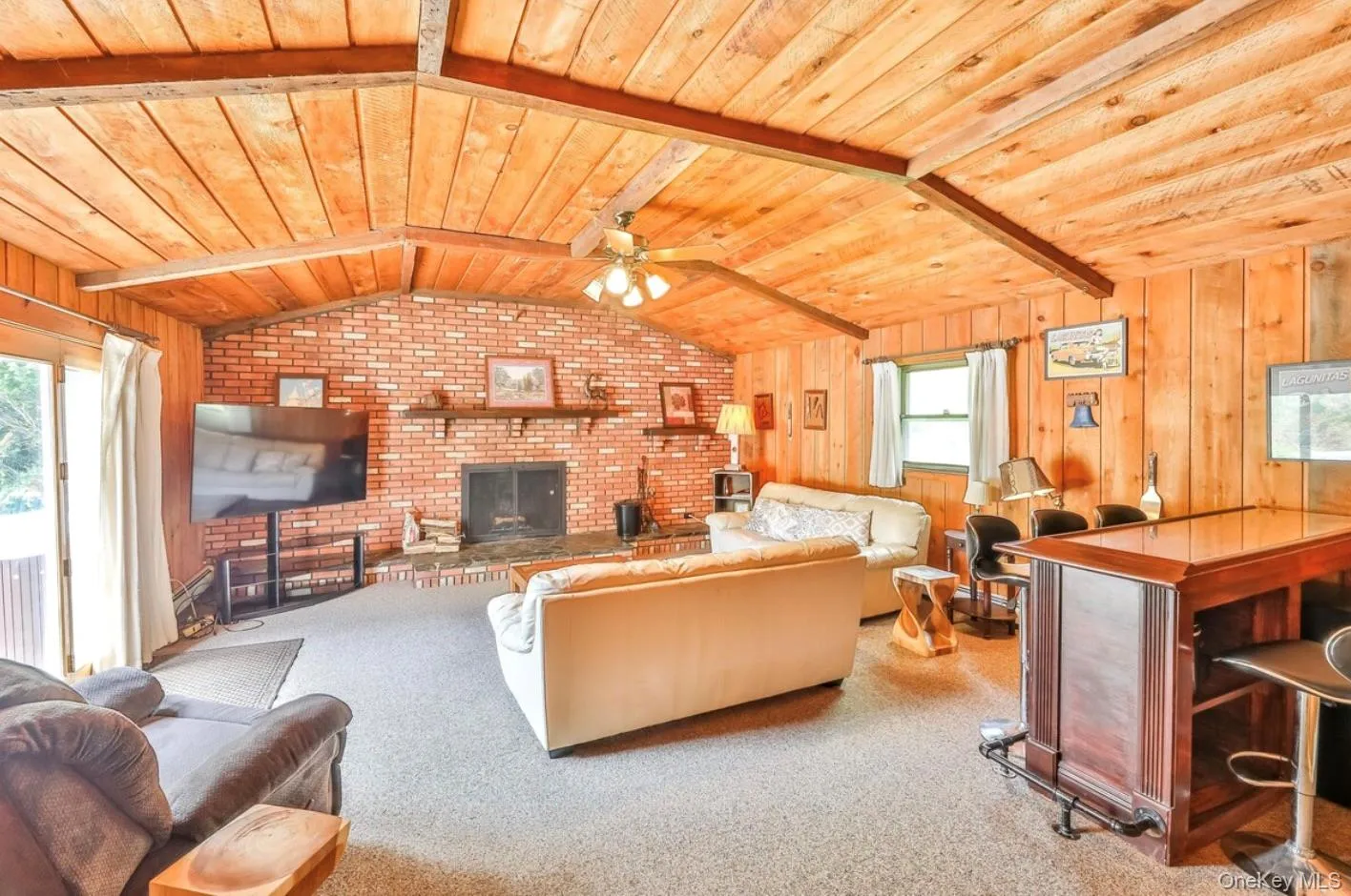 Carpeted living area featuring wooden ceiling, a brick fireplace, wood walls, brick wall, and ceiling fan Carpeted living area featuring wooden ceiling, a brick fireplace, wood walls, brick wall, and ceiling fan