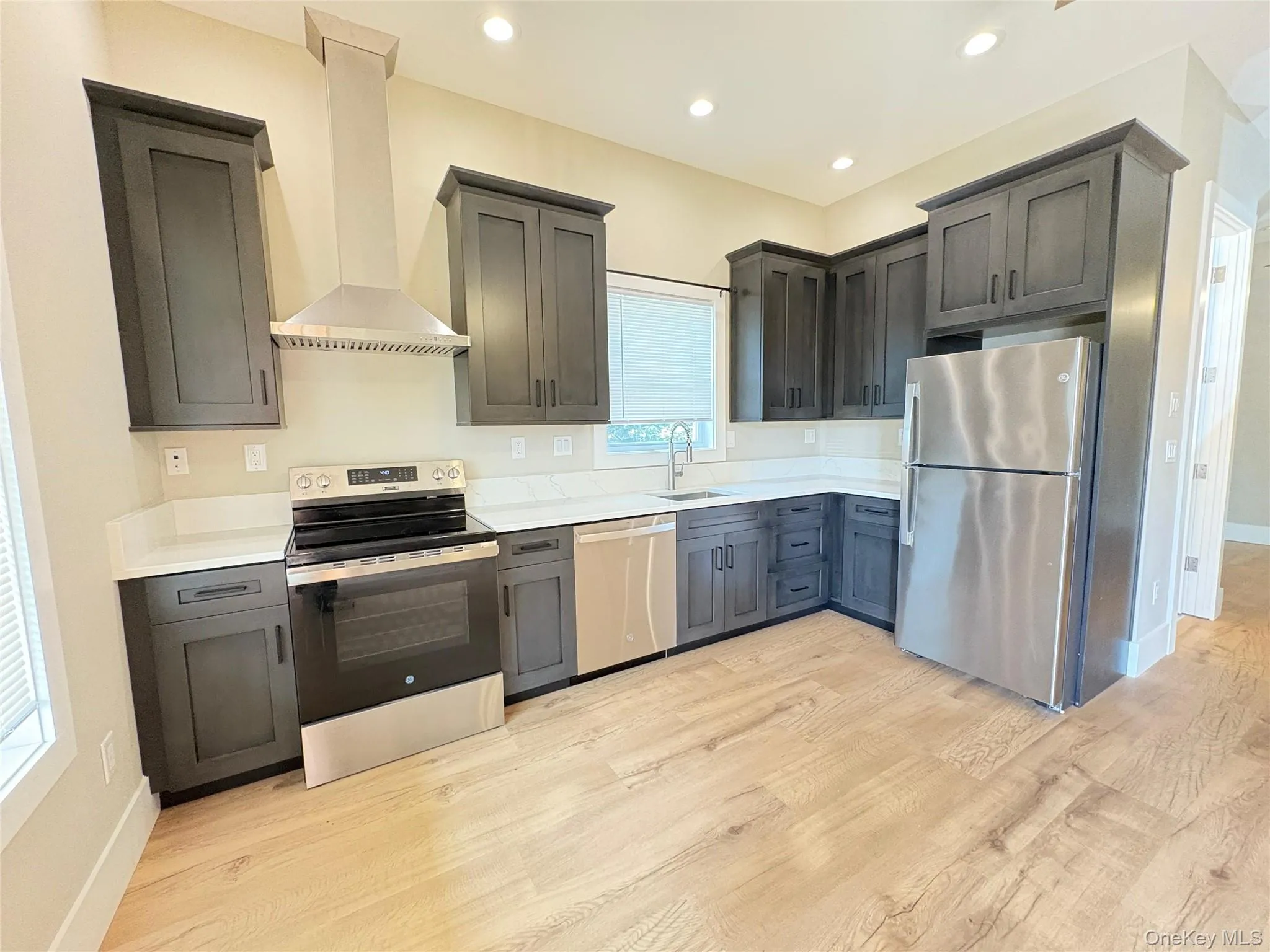 Kitchen with stainless steel appliances, wall chimney exhaust hood, light wood-type flooring, recessed lighting, and light stone counters Kitchen with stainless steel appliances, wall chimney exhaust hood, light wood-type flooring, recessed lighting, and light stone counters