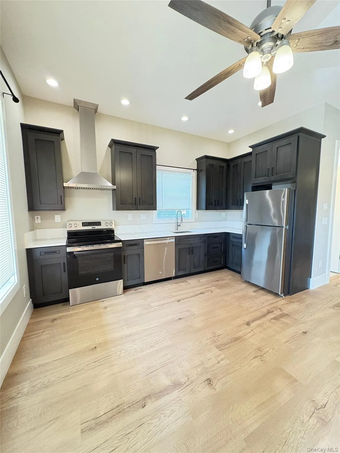 Kitchen featuring stainless steel appliances, wall chimney range hood, light wood finished floors, a ceiling fan, and recessed lighting Kitchen featuring stainless steel appliances, wall chimney range hood, light wood finished floors, a ceiling fan, and recessed lighting