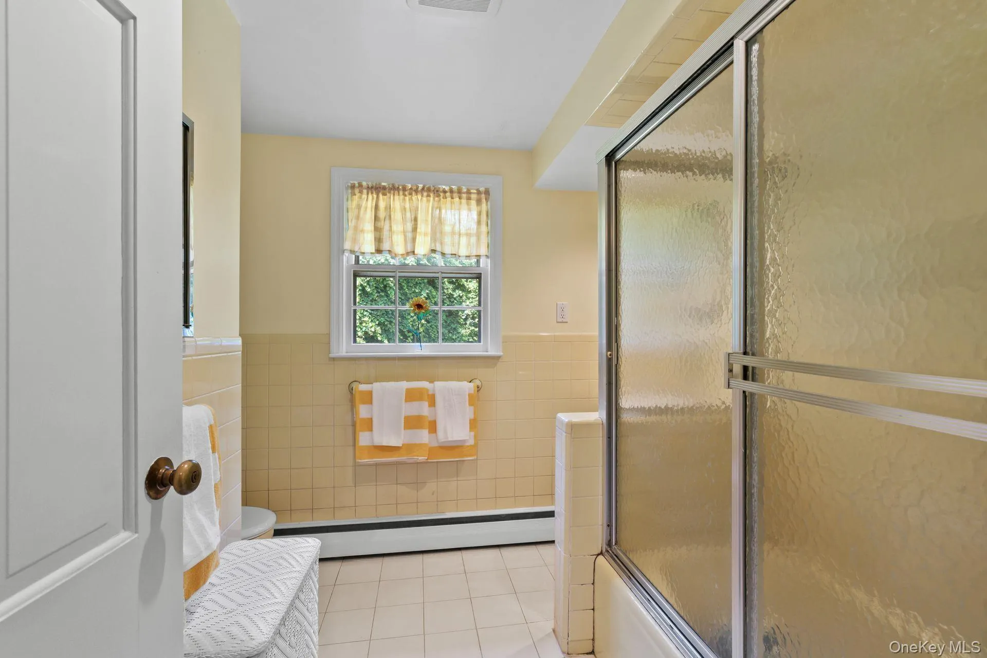 Full bathroom featuring light tile patterned floors, tile walls, wainscoting, a baseboard radiator, and bath / shower combo with glass door Full bathroom featuring light tile patterned floors, tile walls, wainscoting, a baseboard radiator, and bath / shower combo with glass door