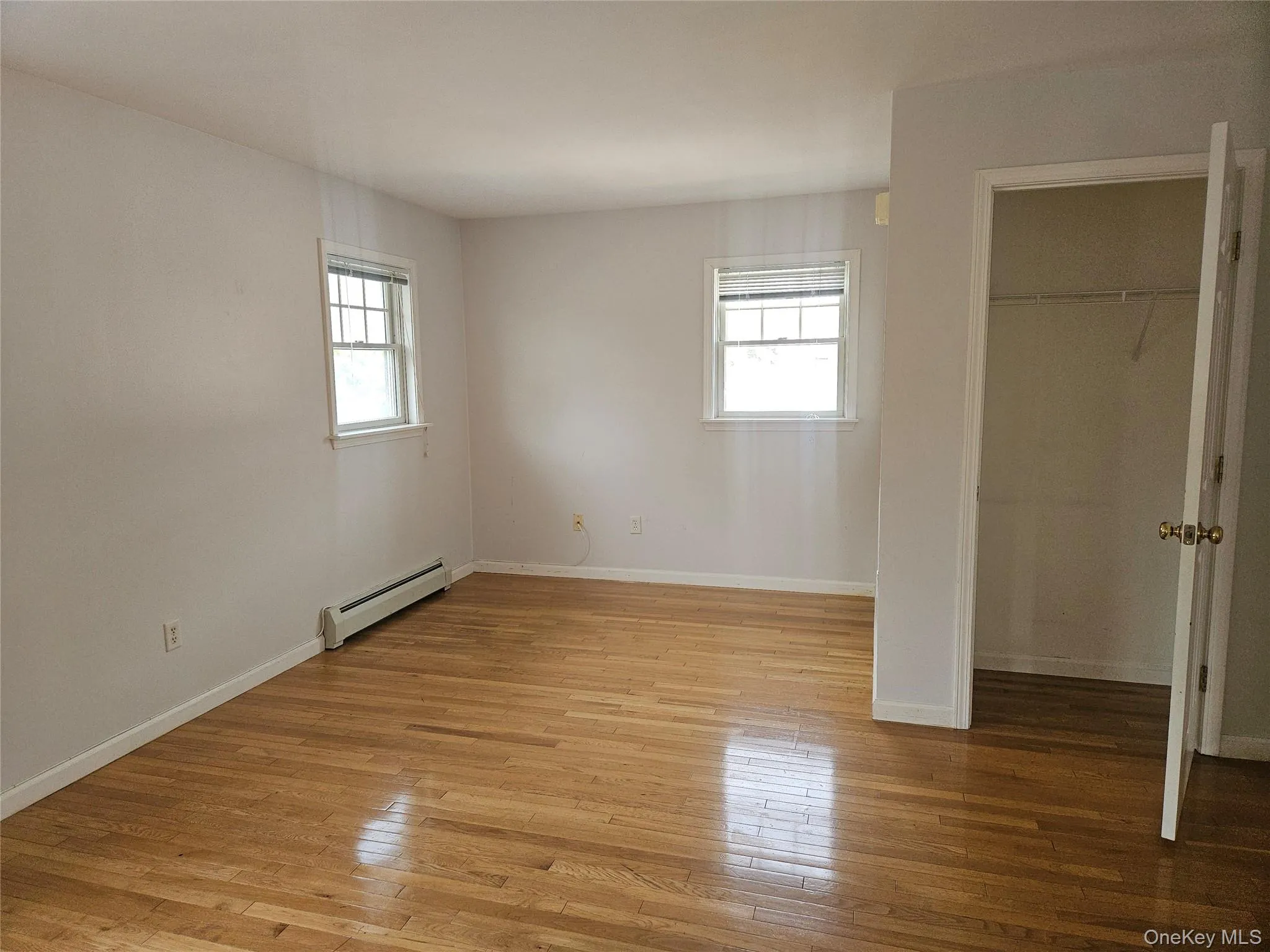 Unfurnished bedroom featuring multiple windows, light wood-style floors, a baseboard radiator, and a spacious closet Unfurnished bedroom featuring multiple windows, light wood-style floors, a baseboard radiator, and a spacious closet
