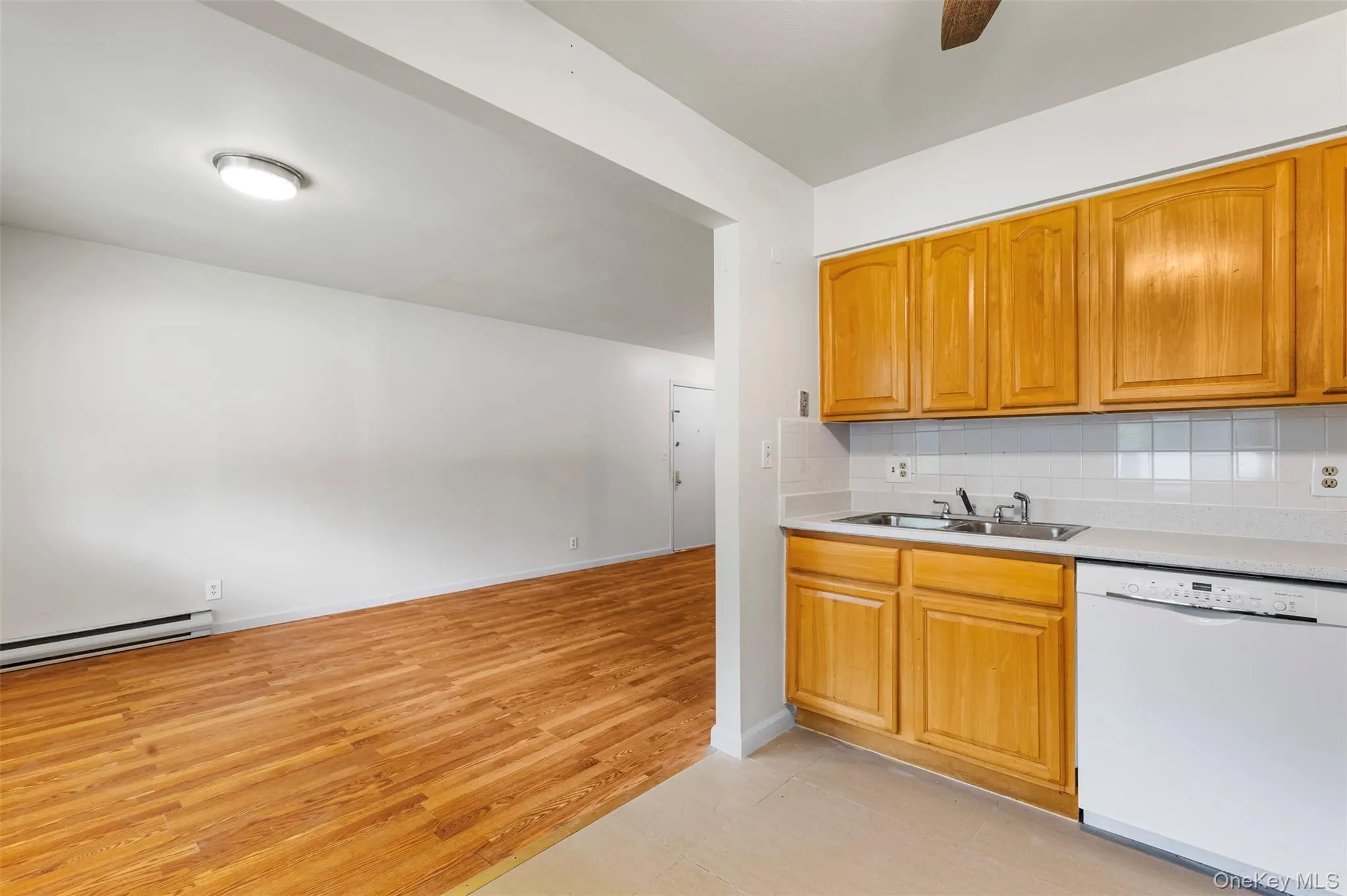 Kitchen featuring white dishwasher, light countertops, backsplash, light wood finished floors, and brown cabinetry Kitchen featuring white dishwasher, light countertops, backsplash, light wood finished floors, and brown cabinetry