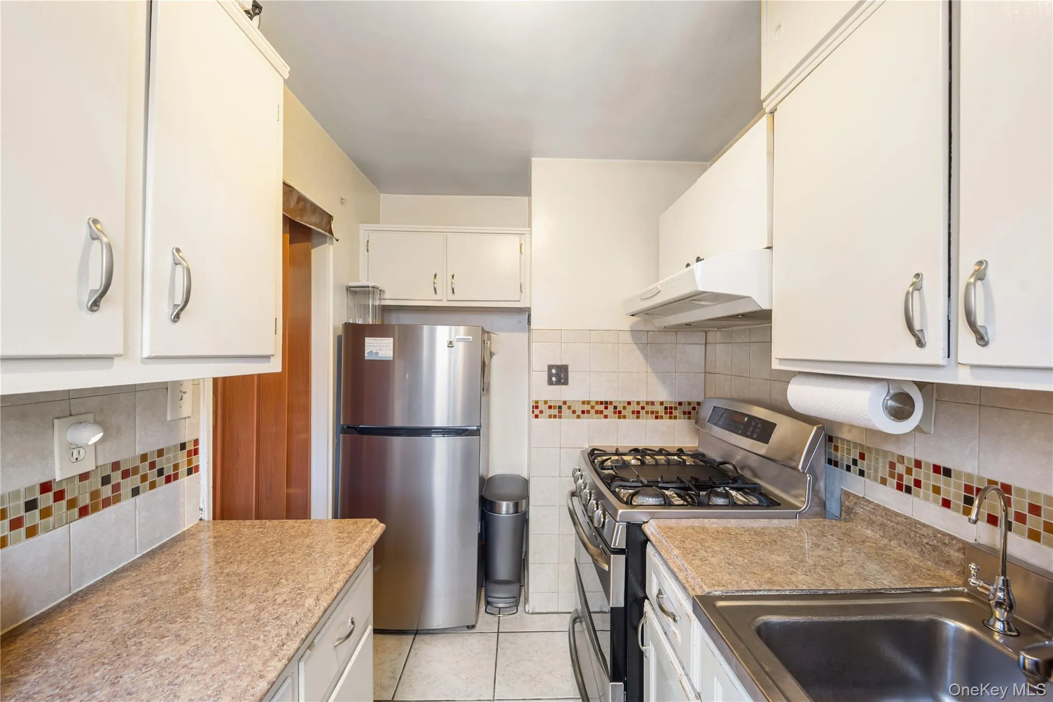 Kitchen with stainless steel appliances, backsplash, light tile patterned floors, under cabinet range hood, and light countertops Kitchen with stainless steel appliances, backsplash, light tile patterned floors, under cabinet range hood, and light countertops