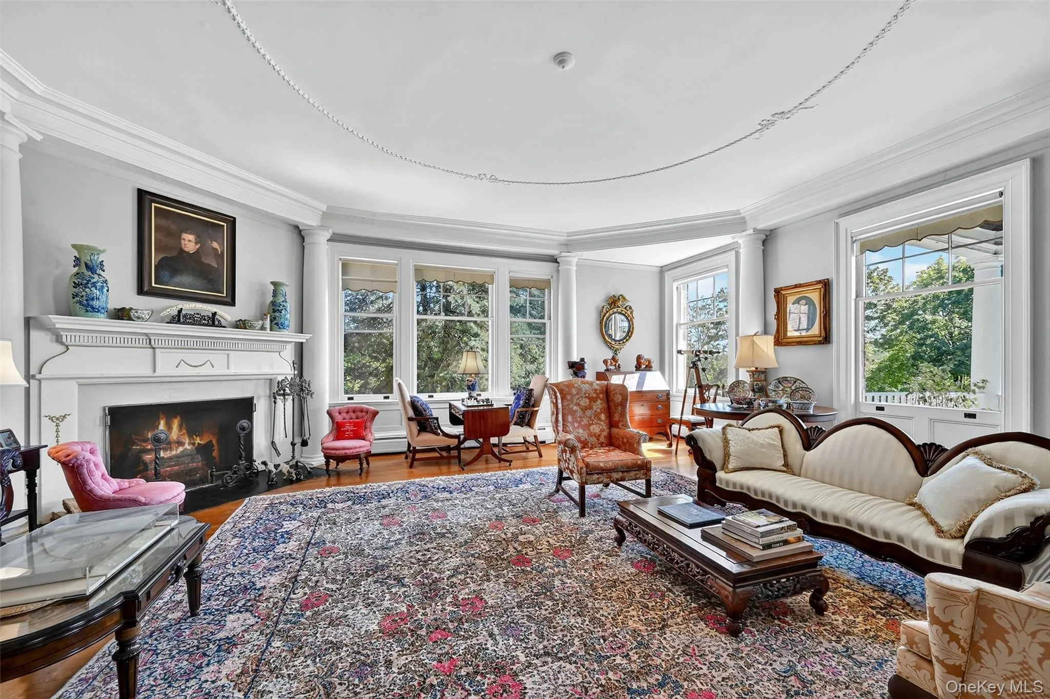 Living room featuring crown molding, wood burning fireplace with millwork surround, ornate columns, and plenty of natural light. Living room featuring crown molding, wood burning fireplace with millwork surround, ornate columns, and plenty of natural light.