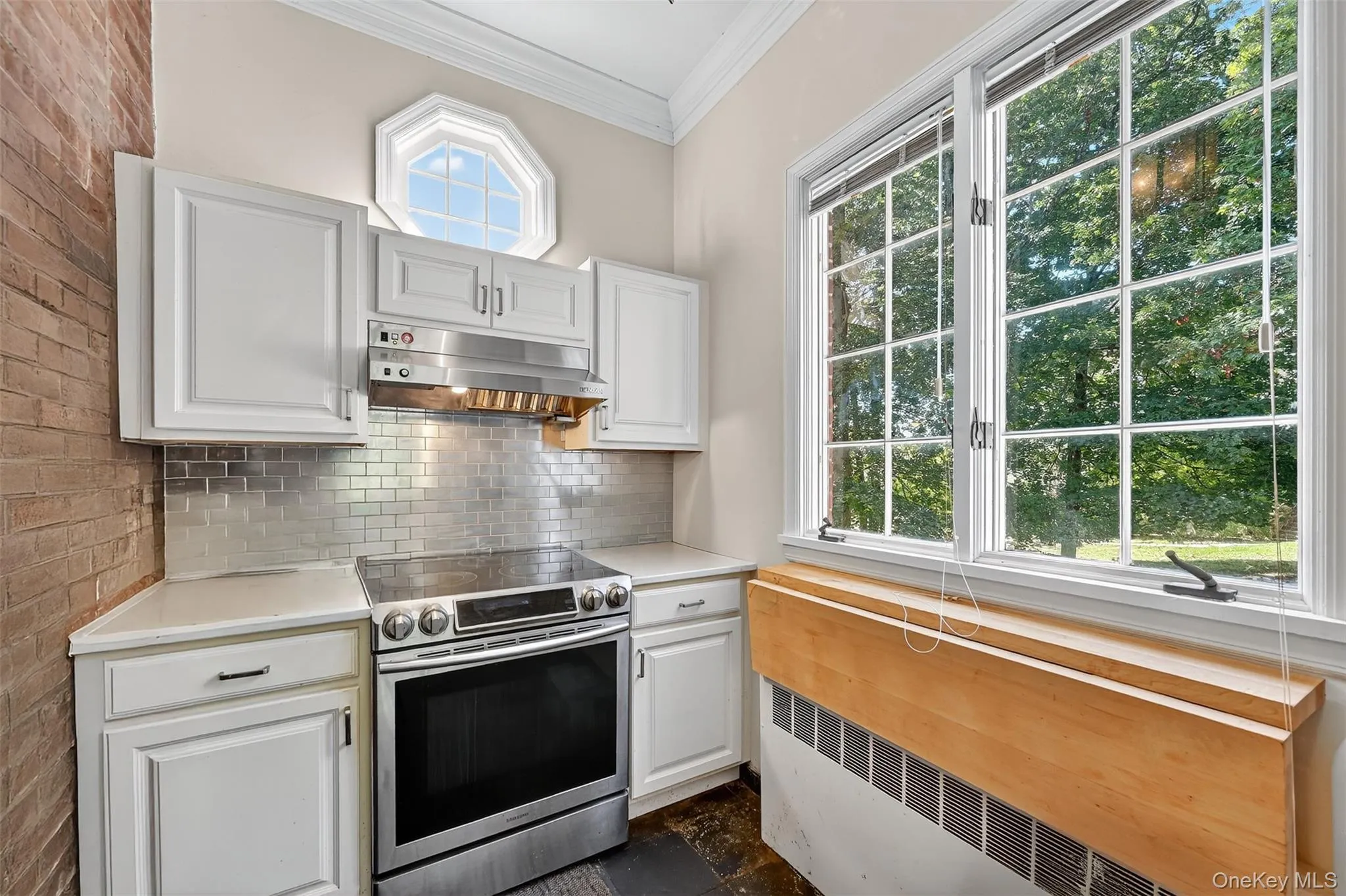 Kitchen with stainless steel electric stove, under cabinet range hood, radiator, ornamental molding, and white cabinetry. Kitchen with stainless steel electric stove, under cabinet range hood, radiator, ornamental molding, and white cabinetry.
