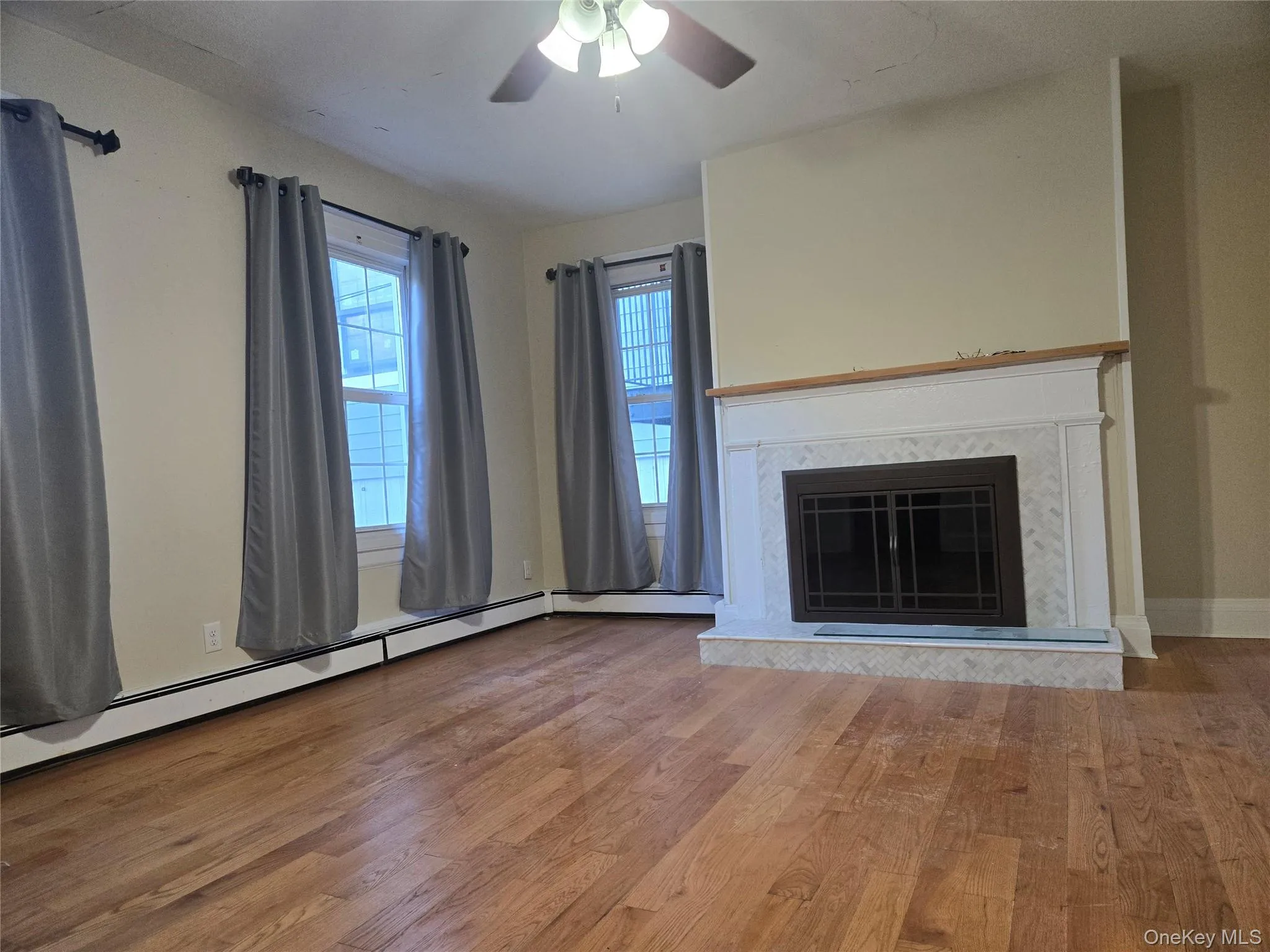 Unfurnished living room with a baseboard radiator, a premium fireplace, wood finished floors, and a ceiling fan Unfurnished living room with a baseboard radiator, a premium fireplace, wood finished floors, and a ceiling fan