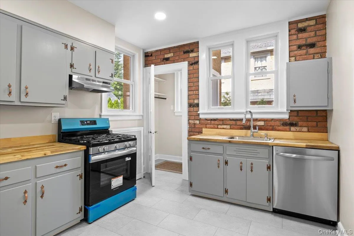 Kitchen featuring gray cabinets, butcher block countertops, appliances with stainless steel finishes, under cabinet range hood, and brick wall Kitchen featuring gray cabinets, butcher block countertops, appliances with stainless steel finishes, under cabinet range hood, and brick wall