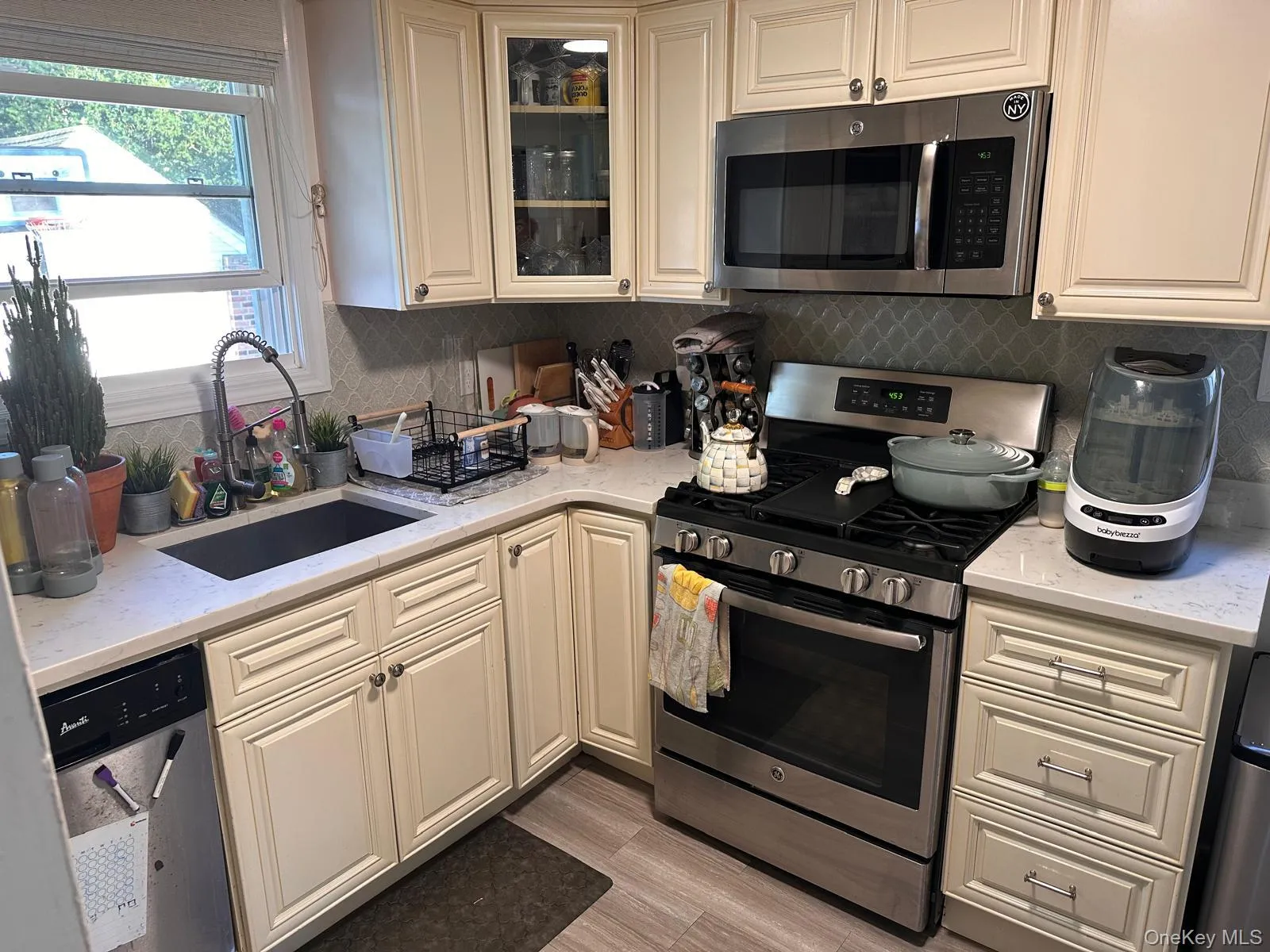 Kitchen featuring appliances with stainless steel finishes, light stone counters, glass insert cabinets, tasteful backsplash, and light wood-style floors Kitchen featuring appliances with stainless steel finishes, light stone counters, glass insert cabinets, tasteful backsplash, and light wood-style floors