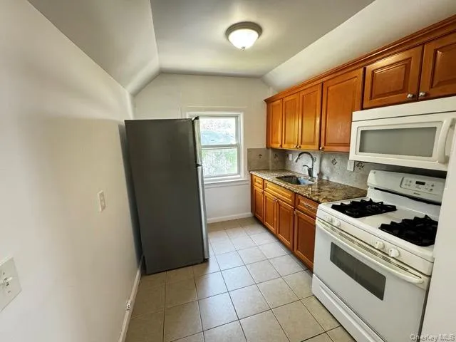Kitchen featuring white appliances, brown cabinets, light tile patterned floors, dark stone counters, and backsplash Kitchen featuring white appliances, brown cabinets, light tile patterned floors, dark stone counters, and backsplash
