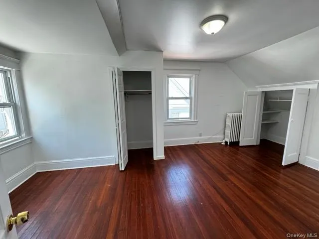 Unfurnished bedroom with multiple closets, dark wood-style flooring, radiator heating unit, and vaulted ceiling Unfurnished bedroom with multiple closets, dark wood-style flooring, radiator heating unit, and vaulted ceiling
