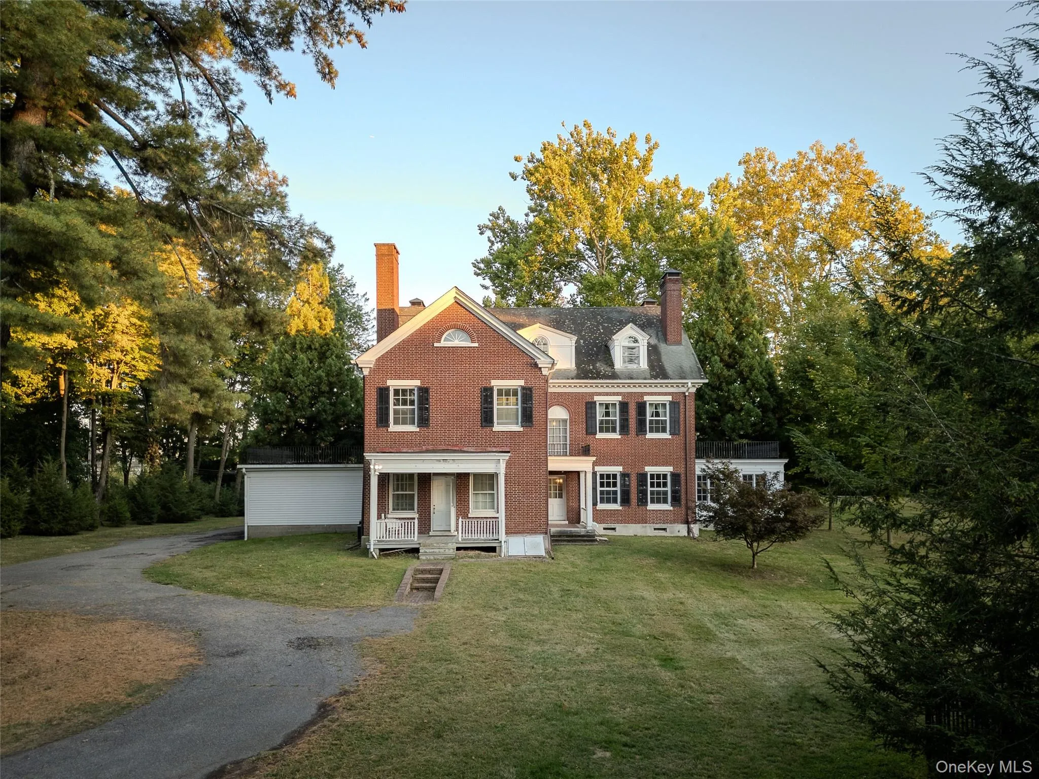 Colonial-style house with a chimney, a front yard, brick siding, driveway, and view of scattered trees Colonial-style house with a chimney, a front yard, brick siding, driveway, and view of scattered trees