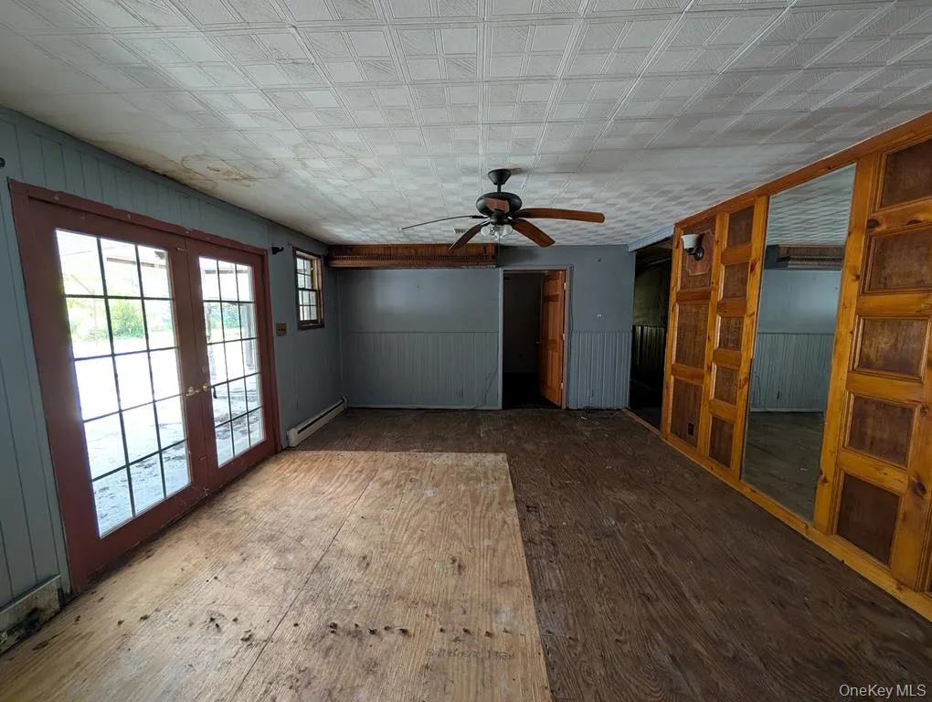 Unfurnished living room with wood-type flooring, french doors, a ceiling fan, wainscoting, and a baseboard radiator Unfurnished living room with wood-type flooring, french doors, a ceiling fan, wainscoting, and a baseboard radiator