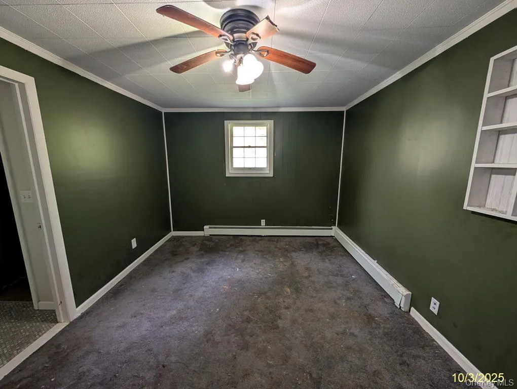 Empty room featuring crown molding, a baseboard radiator, dark colored carpet, and a ceiling fan Empty room featuring crown molding, a baseboard radiator, dark colored carpet, and a ceiling fan