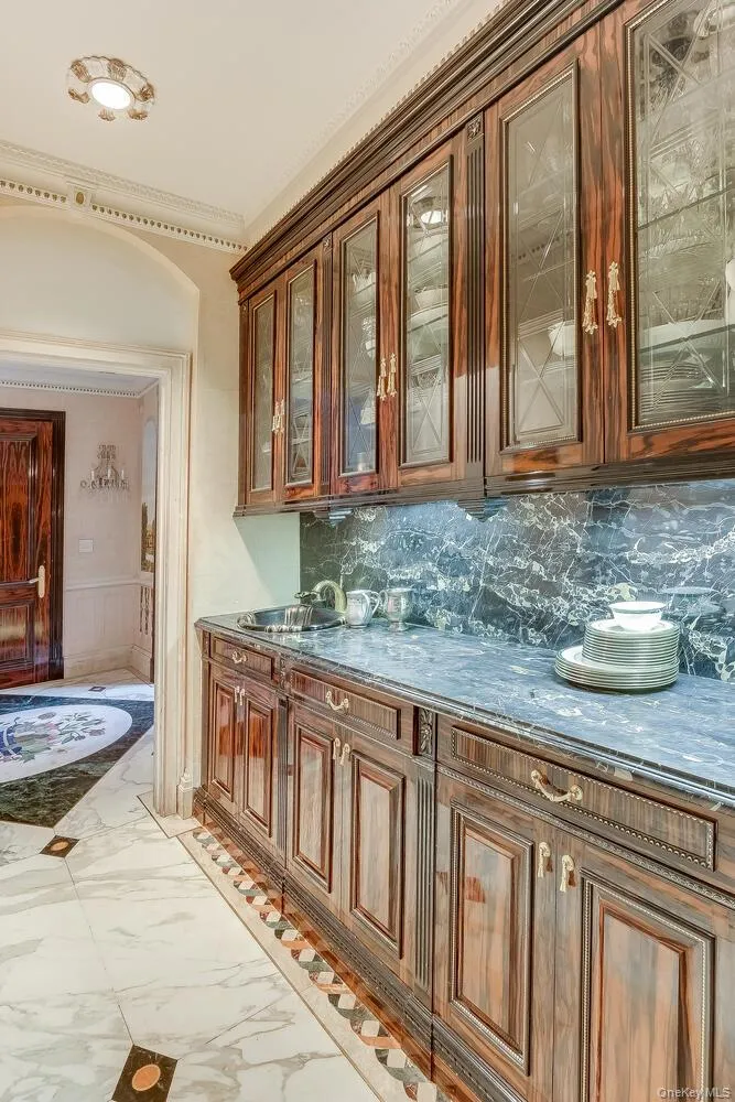 Bar with light marble finish floors, brown cabinetry, glass insert cabinets, decorative backsplash, and dark stone countertops Bar with light marble finish floors, brown cabinetry, glass insert cabinets, decorative backsplash, and dark stone countertops