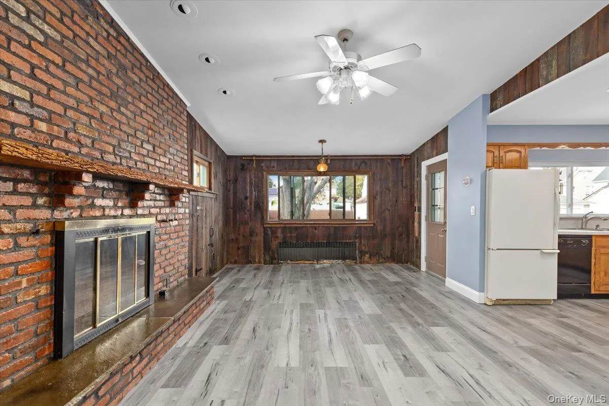 Unfurnished living room featuring wooden walls, light wood-style floors, radiator, a ceiling fan, and a brick fireplace Unfurnished living room featuring wooden walls, light wood-style floors, radiator, a ceiling fan, and a brick fireplace