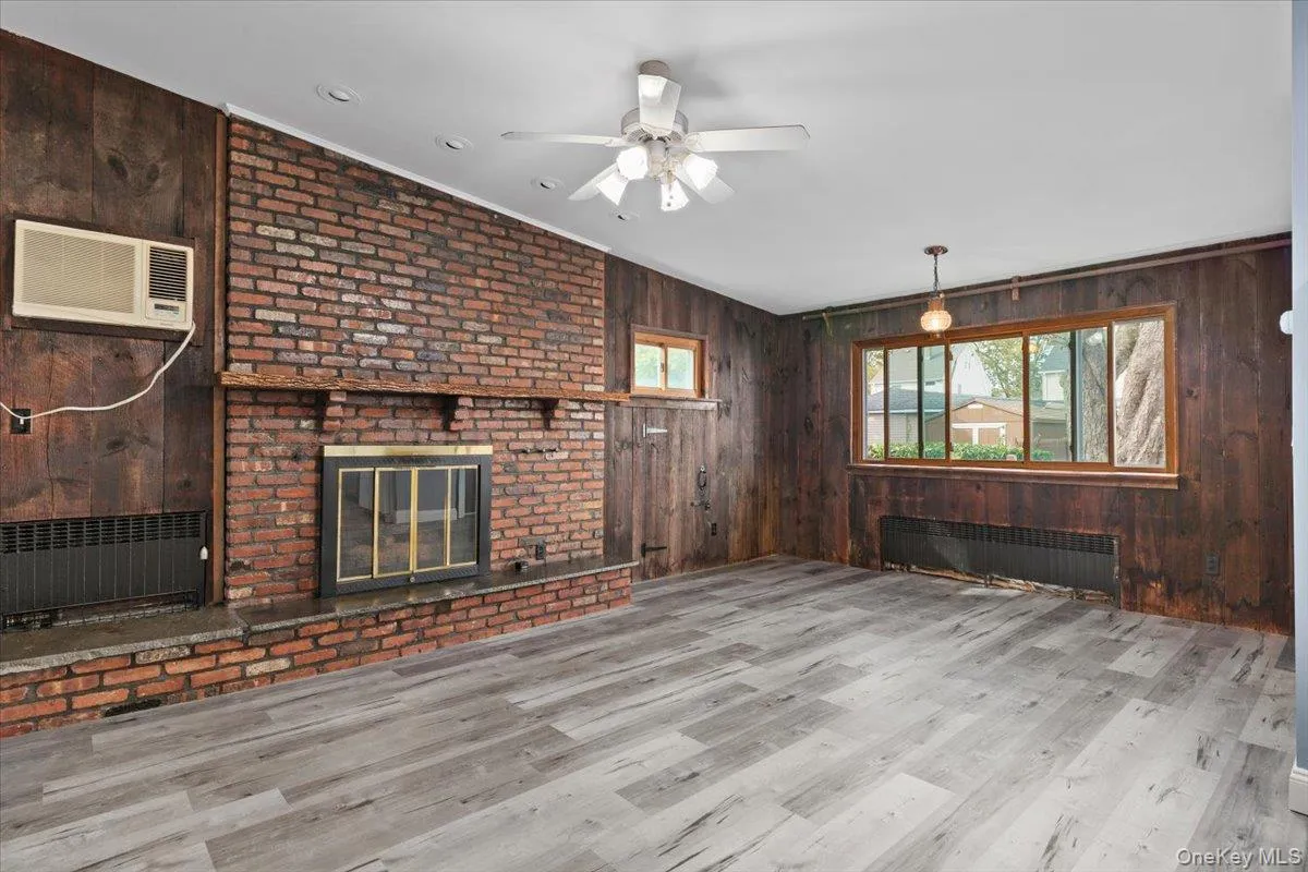 Unfurnished living room featuring a brick fireplace, wood walls, radiator, a wall mounted AC, and crown molding Unfurnished living room featuring a brick fireplace, wood walls, radiator, a wall mounted AC, and crown molding