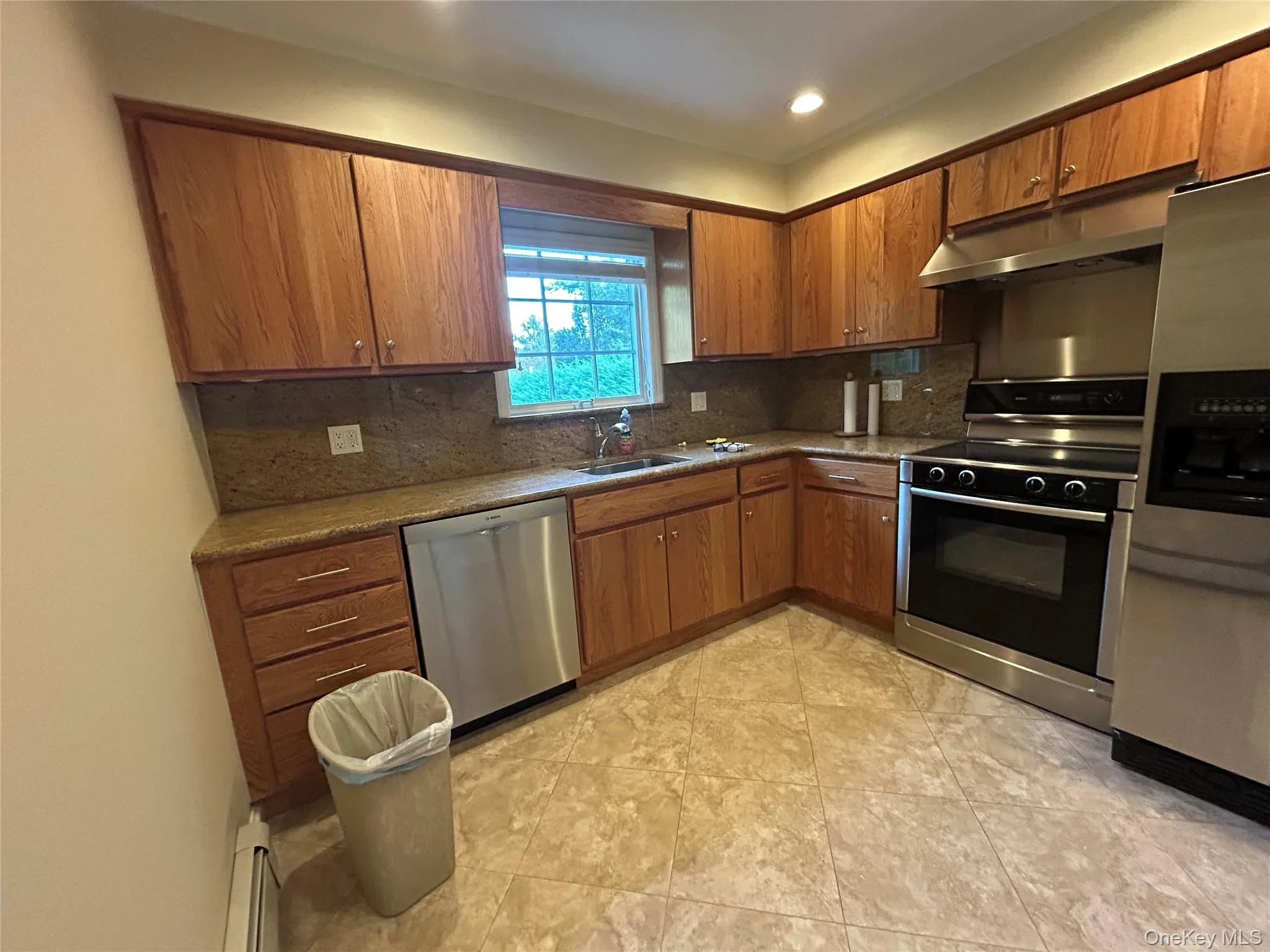 Kitchen with appliances with stainless steel finishes, tasteful backsplash, dark stone counters, brown cabinetry, and recessed lighting Kitchen with appliances with stainless steel finishes, tasteful backsplash, dark stone counters, brown cabinetry, and recessed lighting
