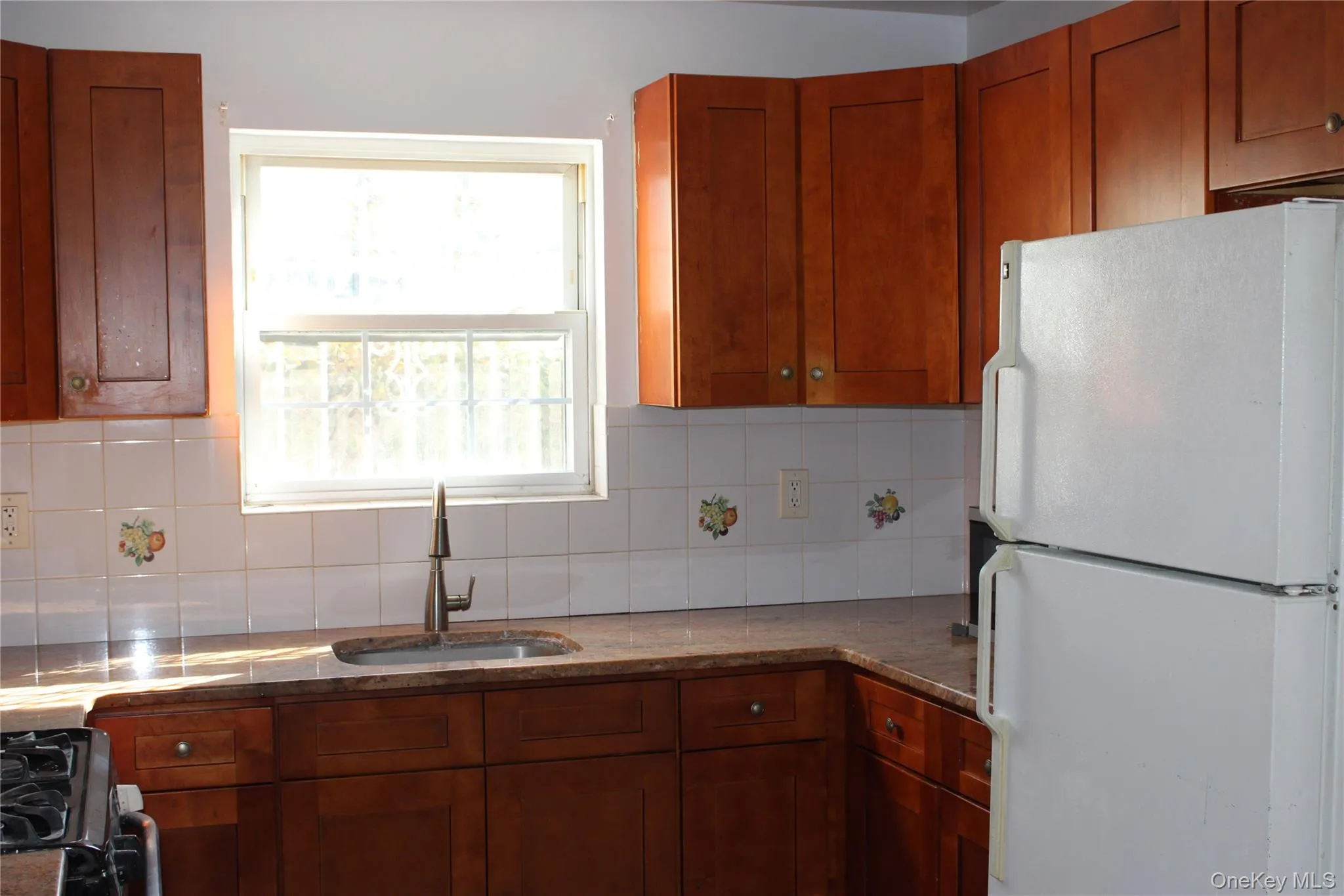 Kitchen with freestanding refrigerator, black range with gas cooktop, brown cabinets, backsplash, and dark stone countertops Kitchen with freestanding refrigerator, black range with gas cooktop, brown cabinets, backsplash, and dark stone countertops