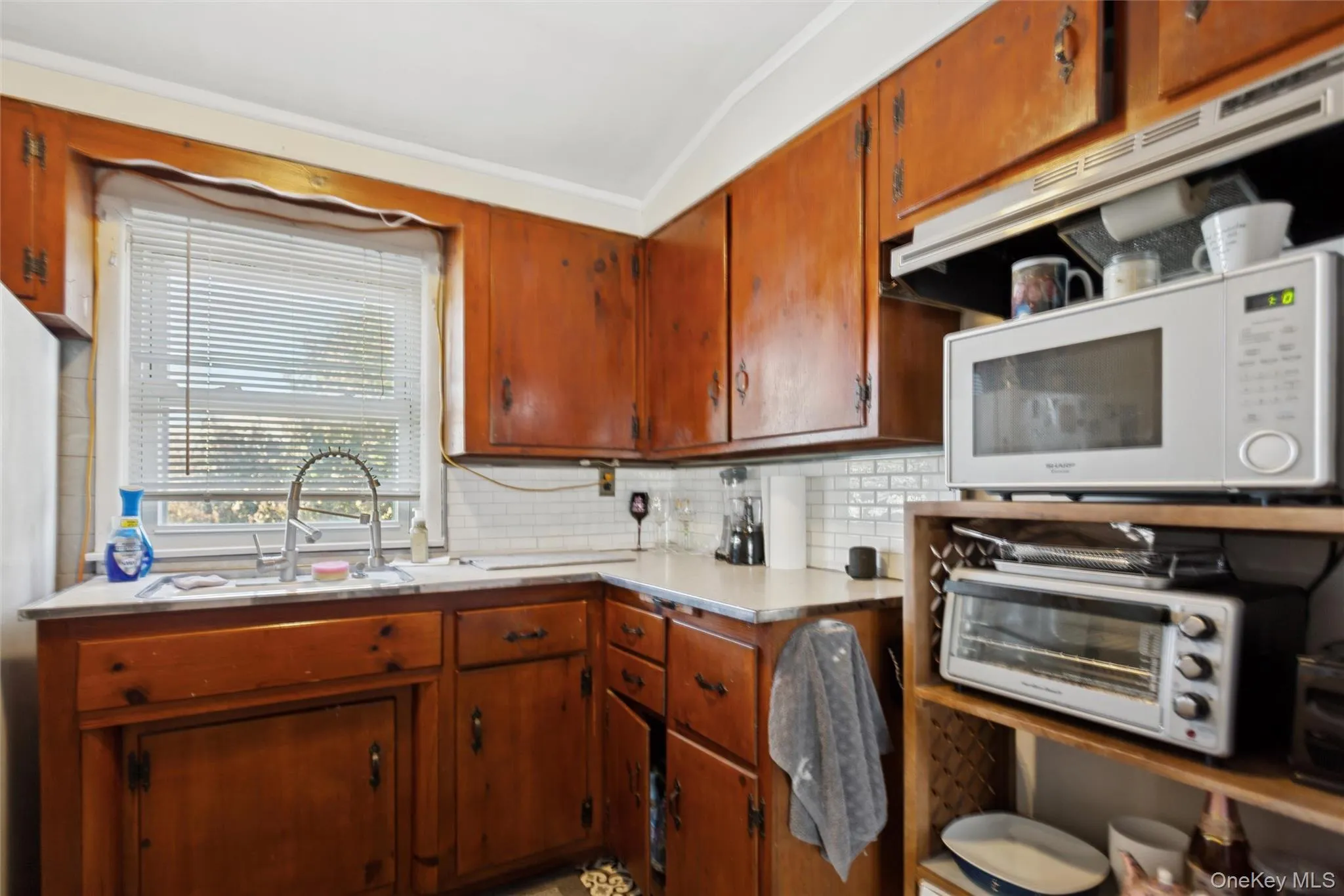 Kitchen featuring light countertops, white microwave, backsplash, brown cabinets, and freestanding refrigerator Kitchen featuring light countertops, white microwave, backsplash, brown cabinets, and freestanding refrigerator