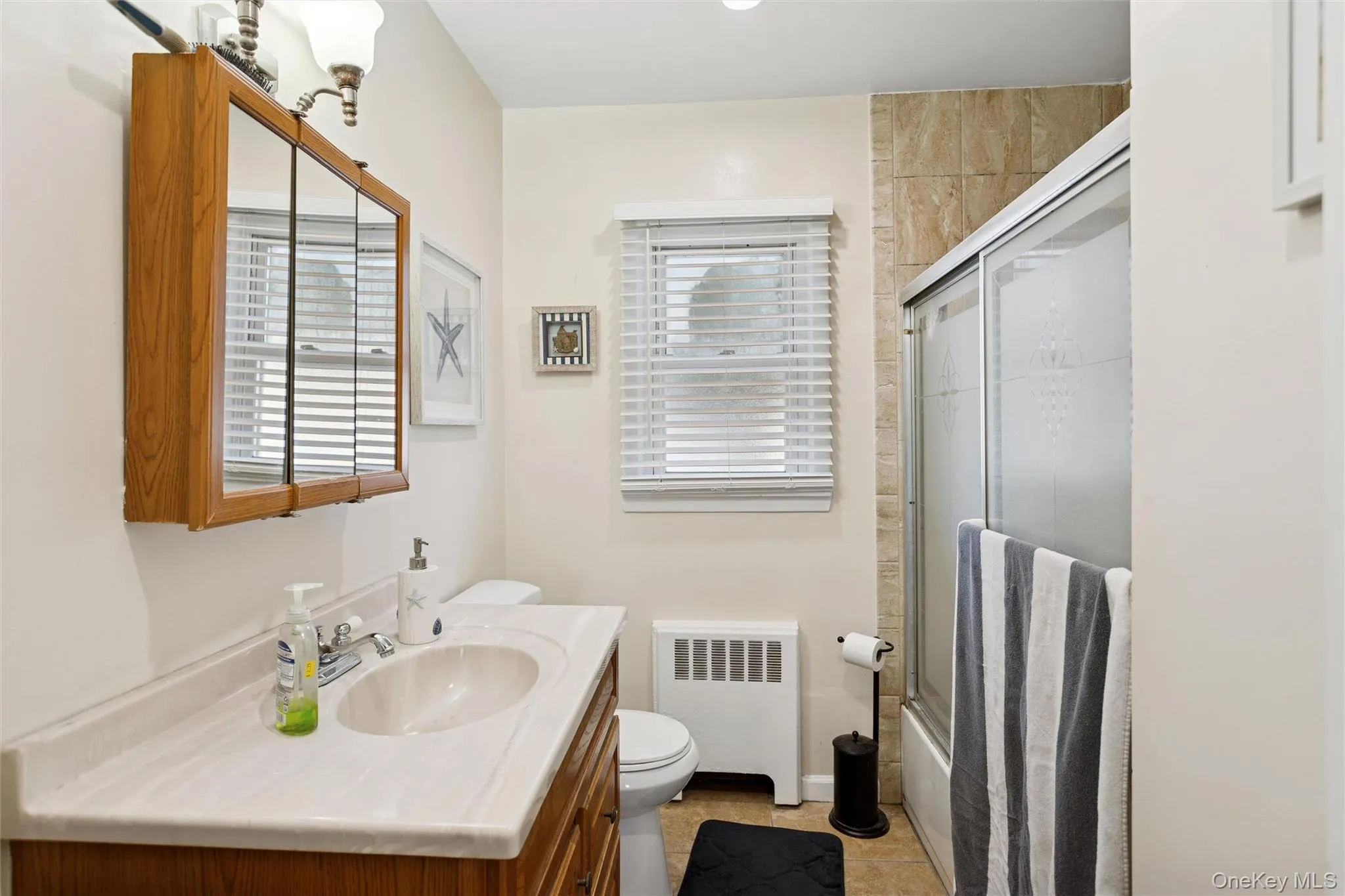 Bathroom with bath / shower combo with glass door, radiator heating unit, vanity, and light tile patterned floors Bathroom with bath / shower combo with glass door, radiator heating unit, vanity, and light tile patterned floors