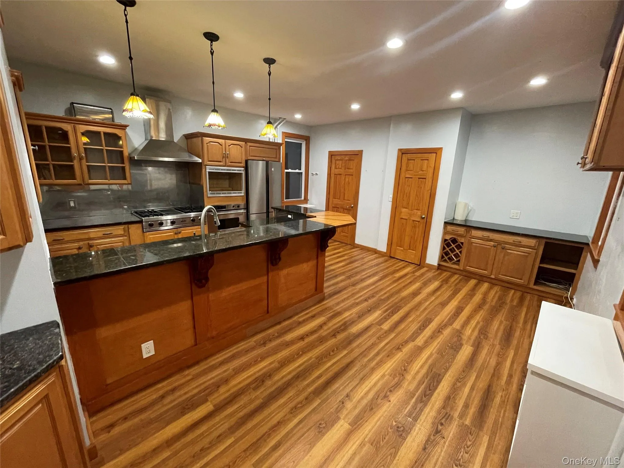 Kitchen with glass insert cabinets, a breakfast bar area, wall chimney range hood, brown cabinets, and recessed lighting Kitchen with glass insert cabinets, a breakfast bar area, wall chimney range hood, brown cabinets, and recessed lighting