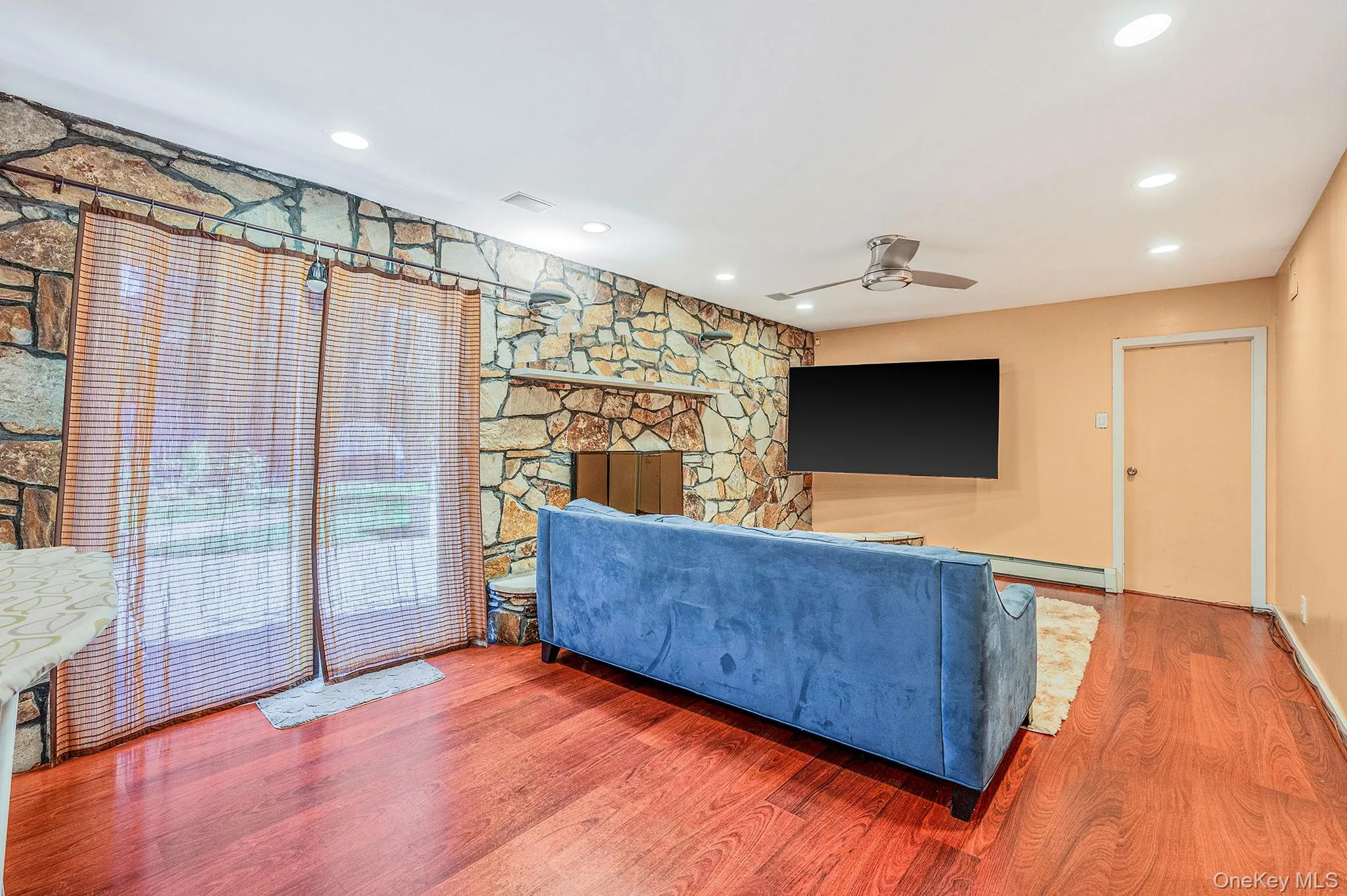 Living room featuring wood finished floors, recessed lighting, ceiling fan, baseboard heating, and a stone fireplace Living room featuring wood finished floors, recessed lighting, ceiling fan, baseboard heating, and a stone fireplace