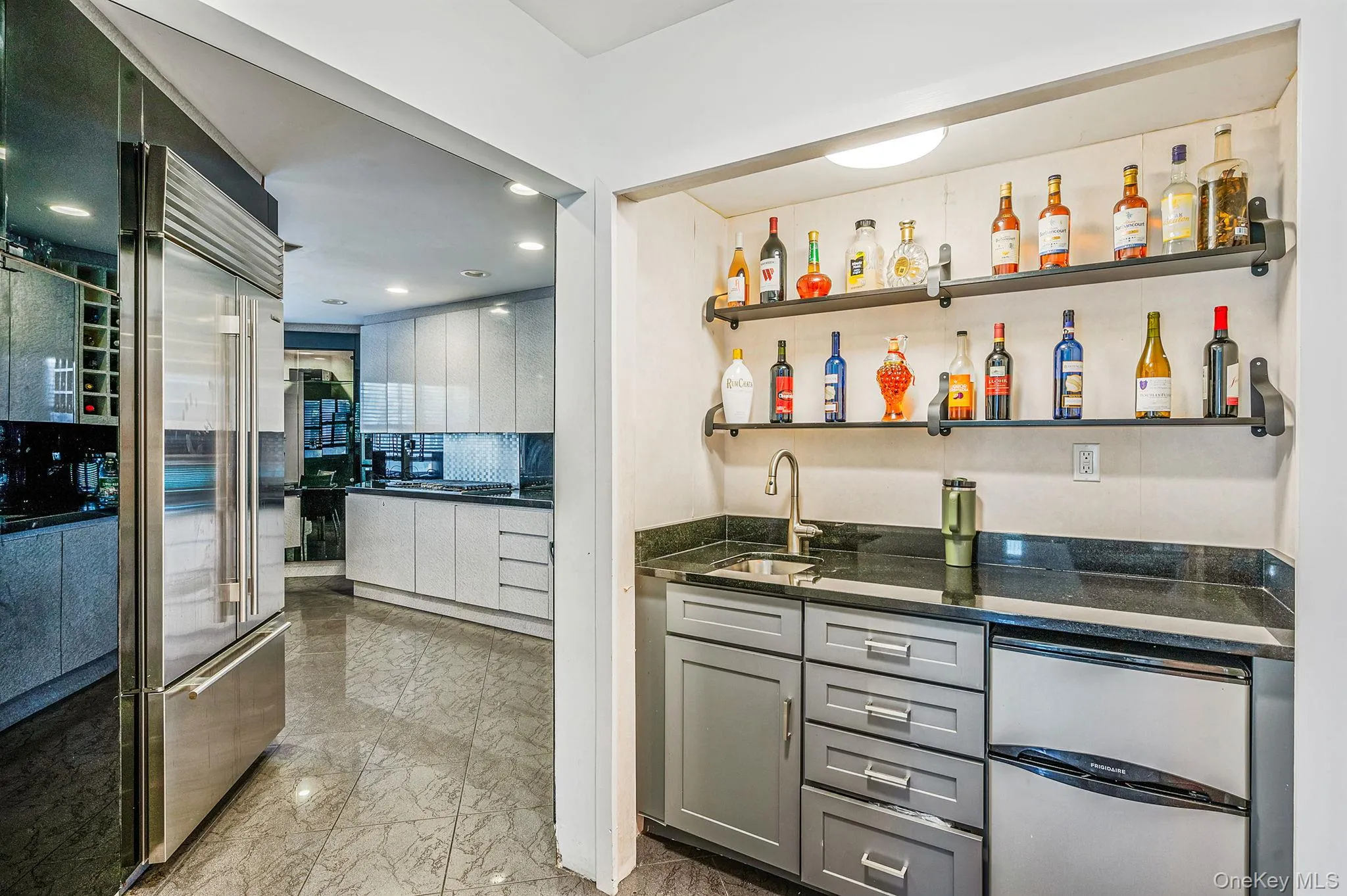 Indoor wet bar with open shelves, light marble finish flooring, gray cabinets, dark stone countertops, and stainless steel refrigerator Indoor wet bar with open shelves, light marble finish flooring, gray cabinets, dark stone countertops, and stainless steel refrigerator