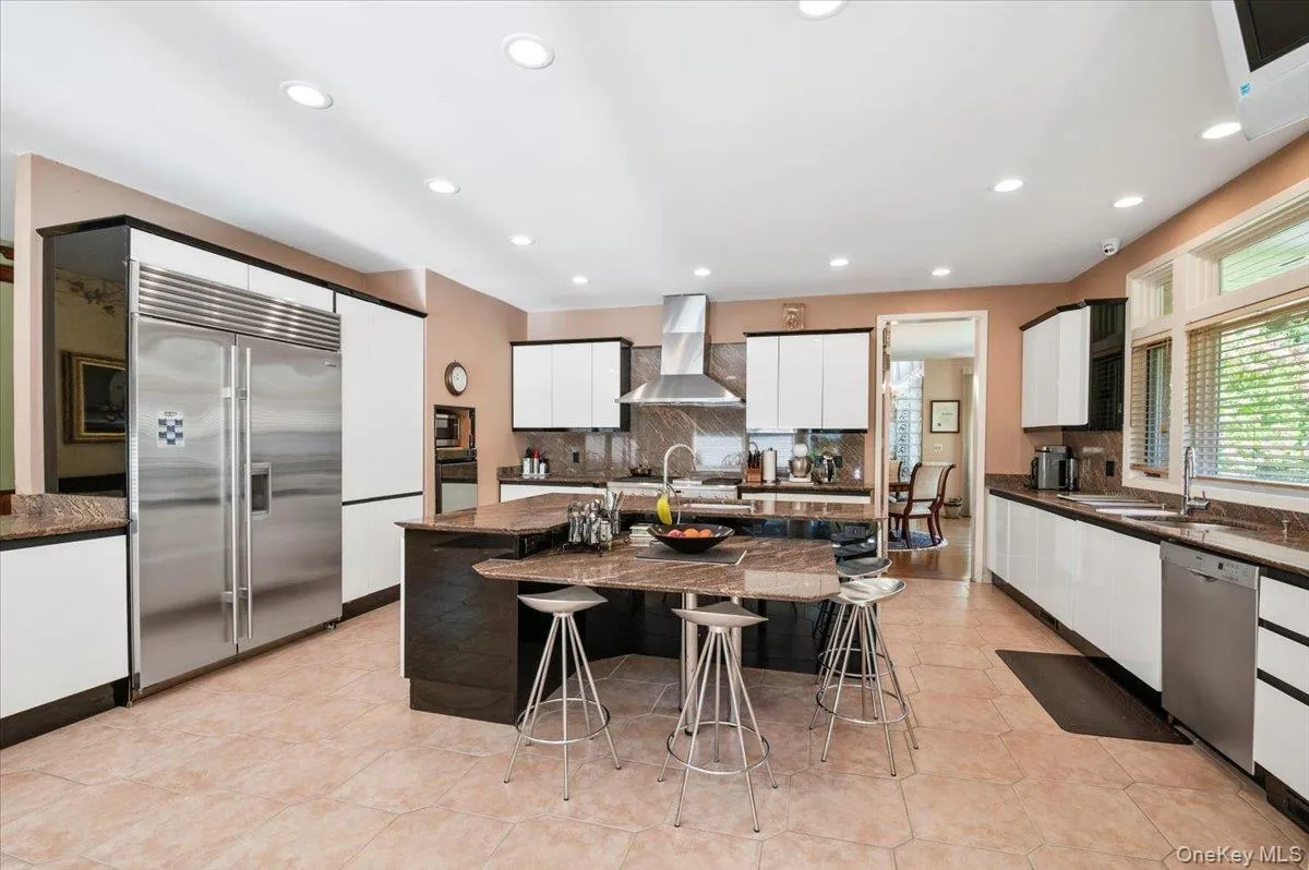 Kitchen with built in appliances, white cabinetry, backsplash, wall chimney range hood, and a center island with sink Kitchen with built in appliances, white cabinetry, backsplash, wall chimney range hood, and a center island with sink