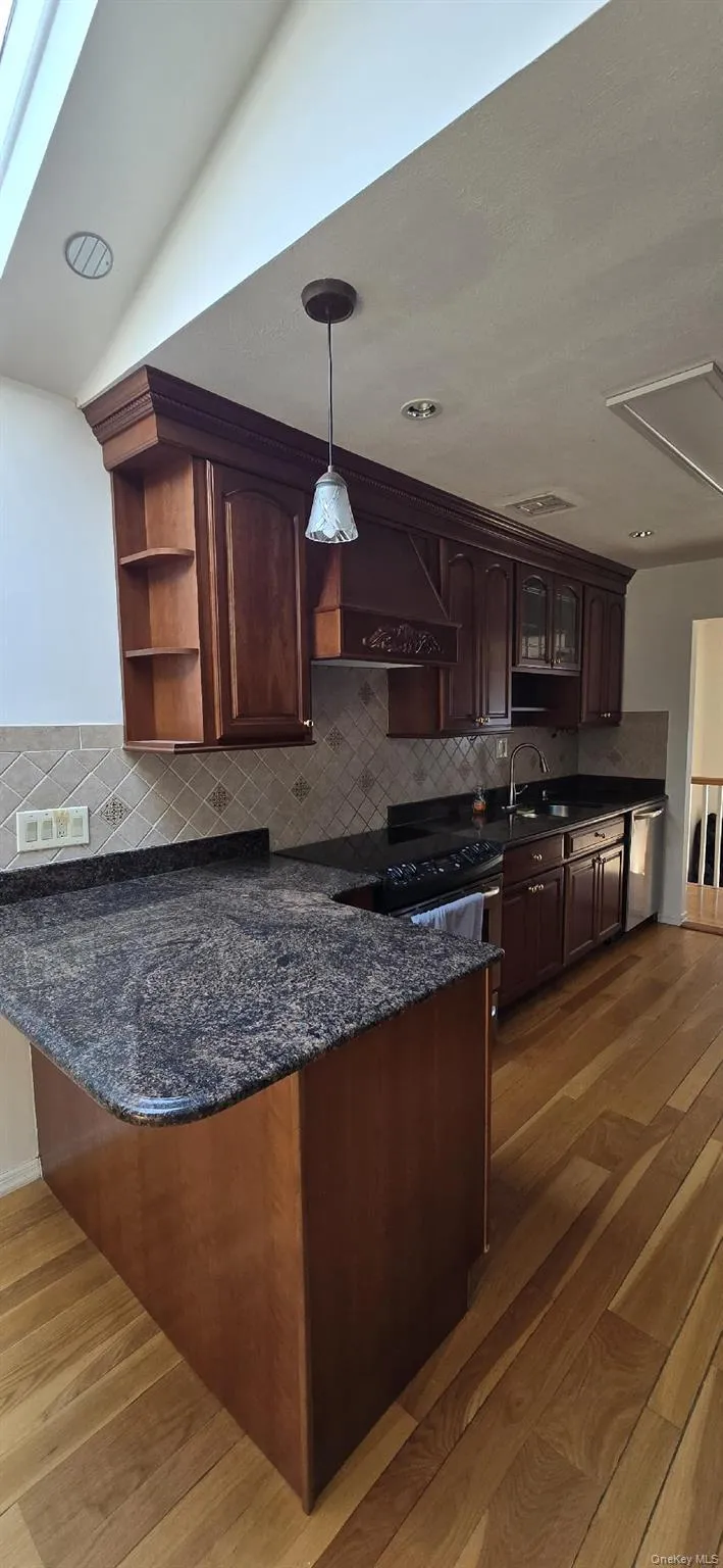 Kitchen featuring rich wood cabinetry, patterned tile backsplash, dark stone countertops, stainless steel appliances, and wood-finish flooring Kitchen featuring rich wood cabinetry, patterned tile backsplash, dark stone countertops, stainless steel appliances, and wood-finish flooring