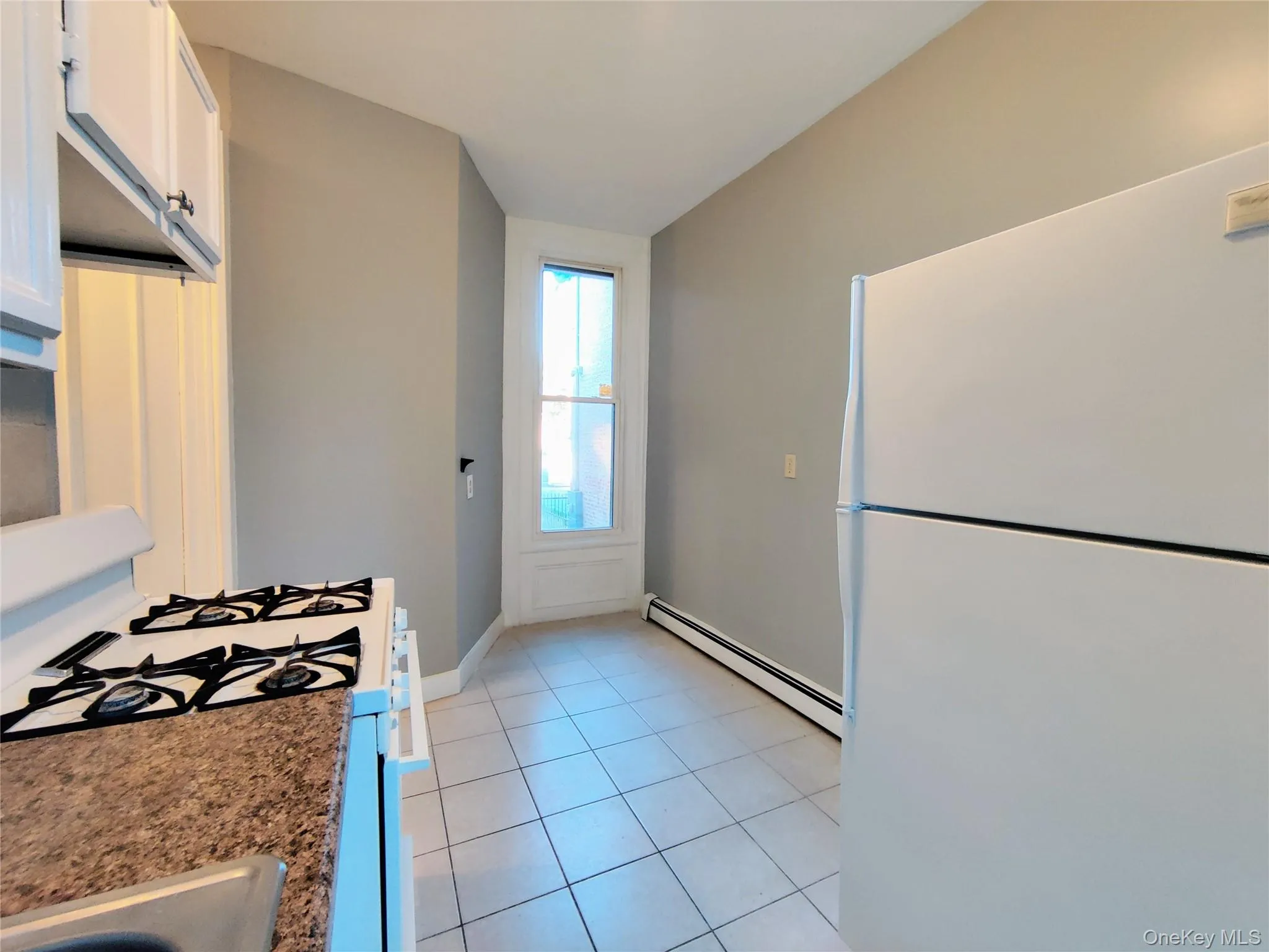 Kitchen with freestanding refrigerator, gas range, baseboard heating, light tile patterned flooring, and white cabinetry Kitchen with freestanding refrigerator, gas range, baseboard heating, light tile patterned flooring, and white cabinetry