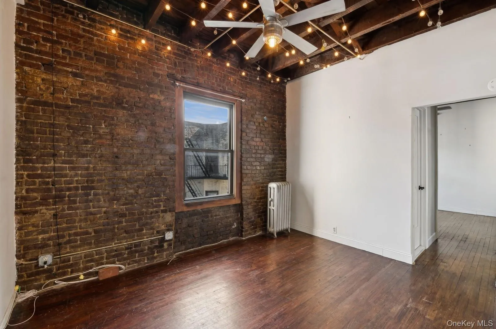 Empty room with wood-type flooring, brick wall, ceiling fan, beam ceiling, and radiator heating unit Empty room with wood-type flooring, brick wall, ceiling fan, beam ceiling, and radiator heating unit