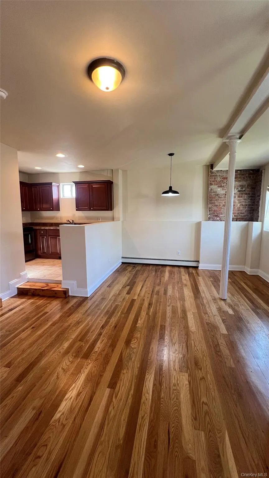 Unfurnished living room featuring light wood-style floors, a baseboard heating unit, and brick wall Unfurnished living room featuring light wood-style floors, a baseboard heating unit, and brick wall