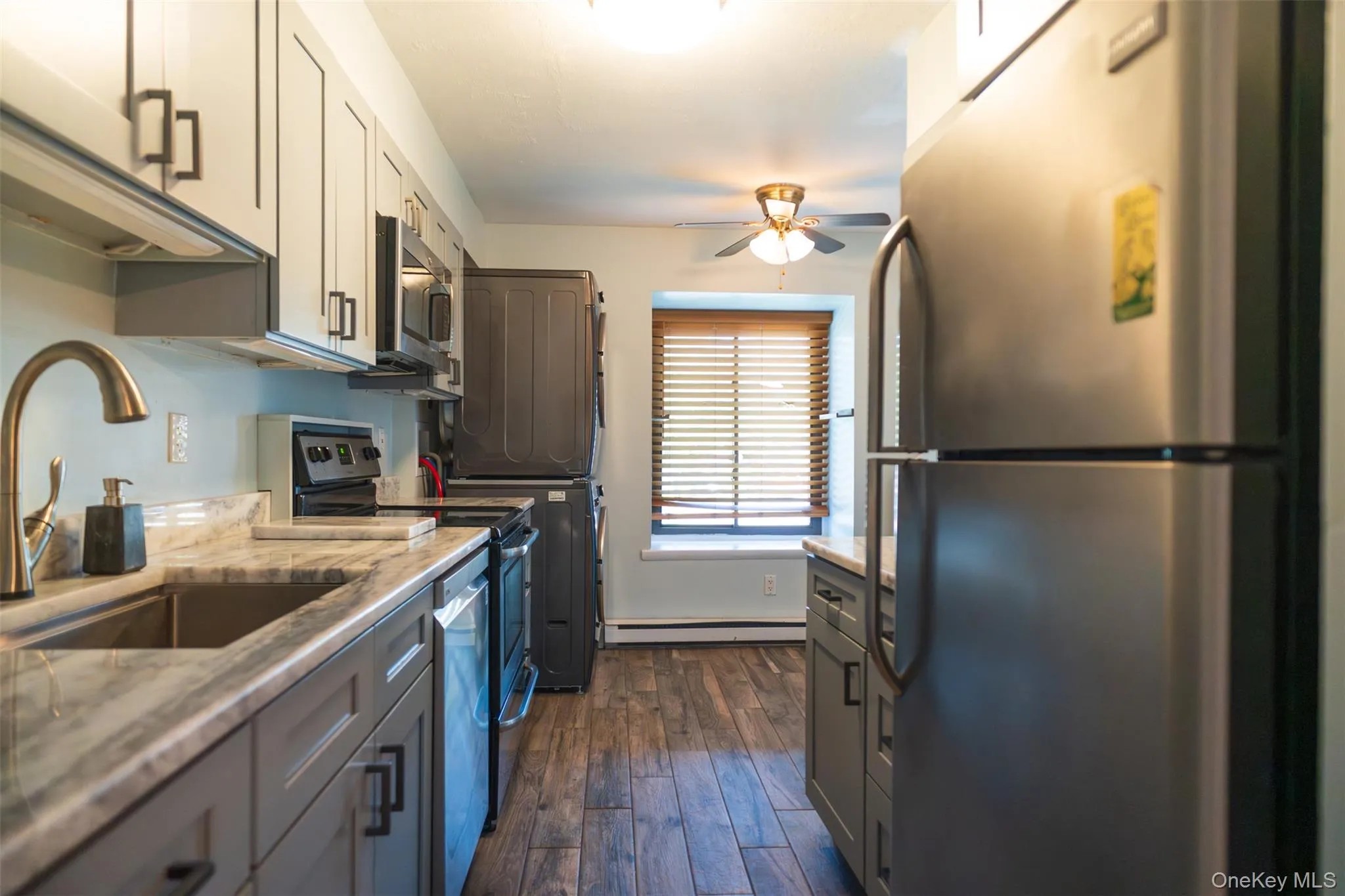 Kitchen with appliances with stainless steel finishes, dark wood finished floors, ceiling fan, light stone countertops, and baseboard heating Kitchen with appliances with stainless steel finishes, dark wood finished floors, ceiling fan, light stone countertops, and baseboard heating
