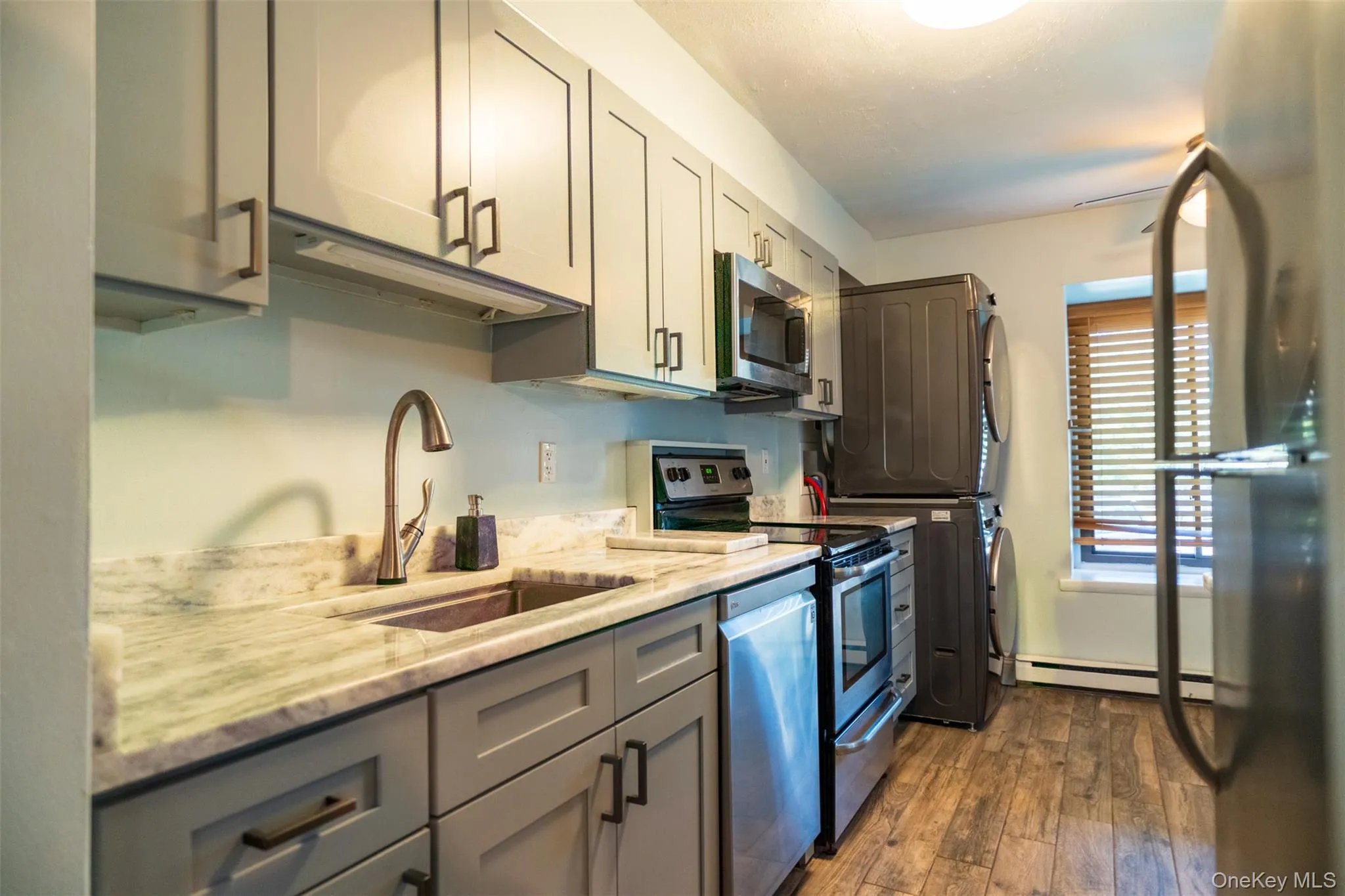 Kitchen featuring gray cabinets, appliances with stainless steel finishes, dark wood-style floors, stacked washer / drying machine, and light stone counters Kitchen featuring gray cabinets, appliances with stainless steel finishes, dark wood-style floors, stacked washer / drying machine, and light stone counters