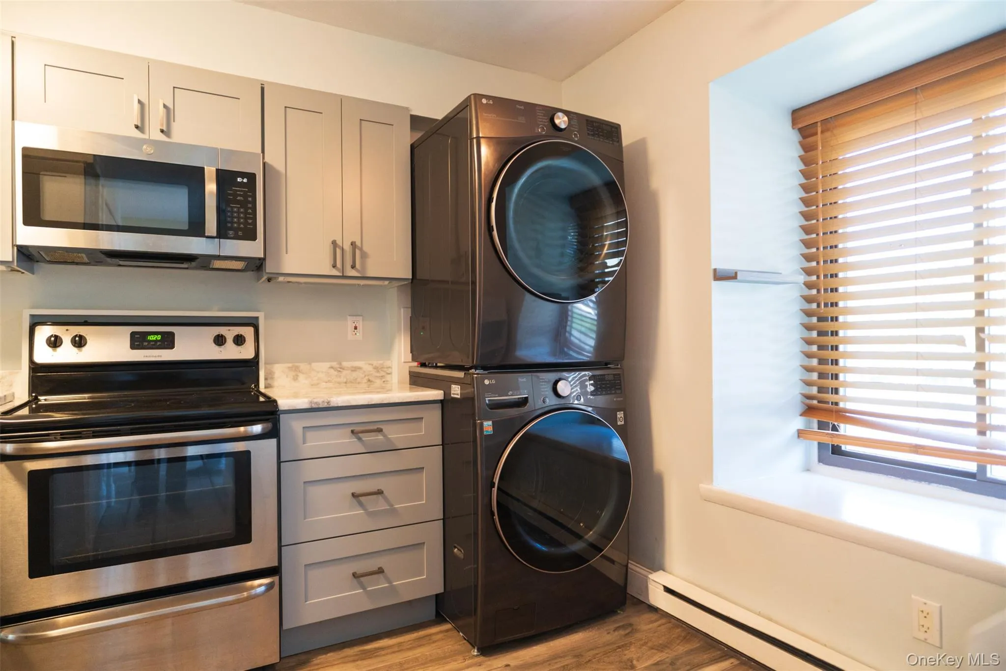 Kitchen with gray cabinets, stainless steel appliances, a baseboard radiator, light wood-type flooring, and stacked washing machine and dryer Kitchen with gray cabinets, stainless steel appliances, a baseboard radiator, light wood-type flooring, and stacked washing machine and dryer