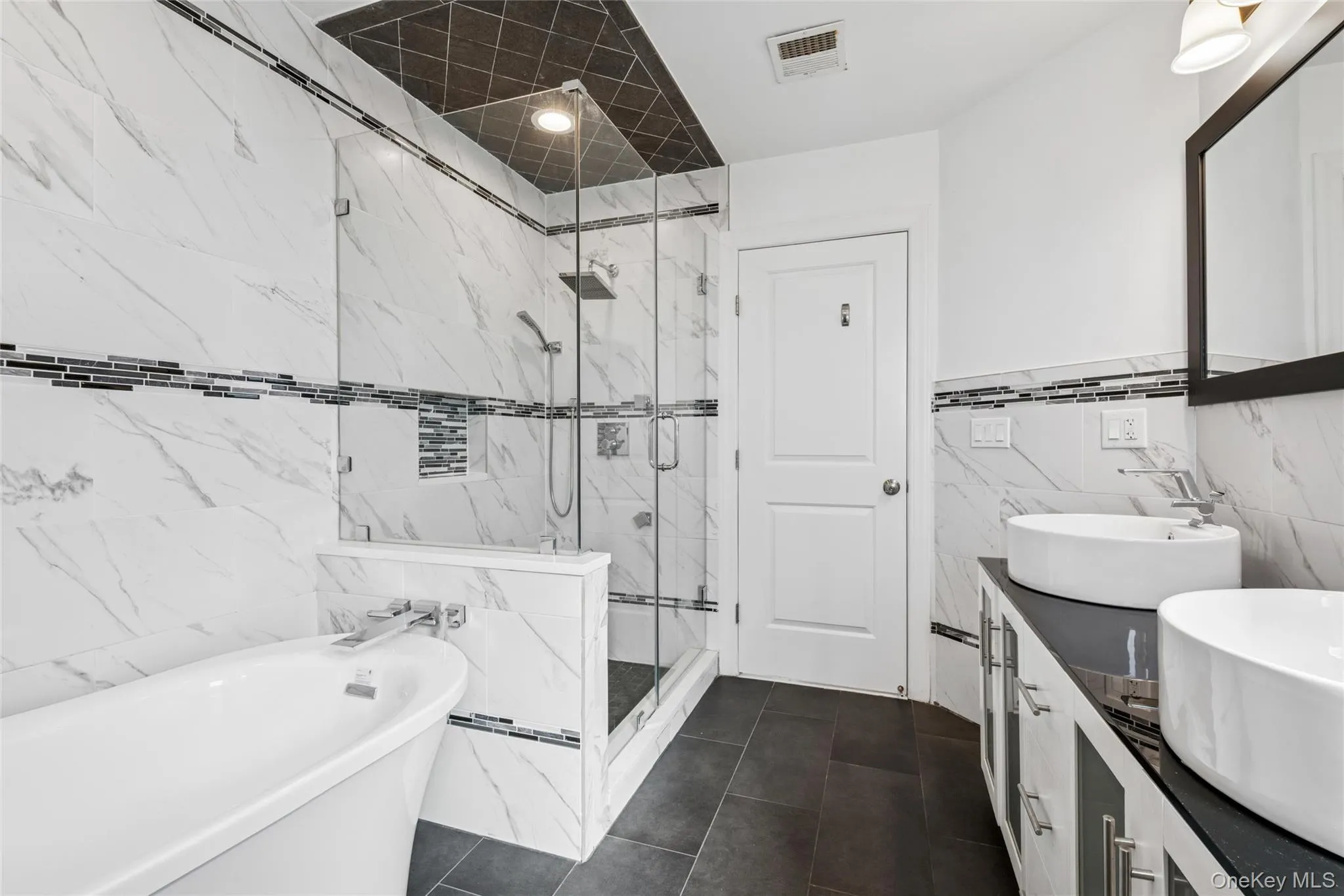 Bathroom with tile walls, a marble finish shower, a freestanding bath, double vanity, and dark tile patterned floors Bathroom with tile walls, a marble finish shower, a freestanding bath, double vanity, and dark tile patterned floors