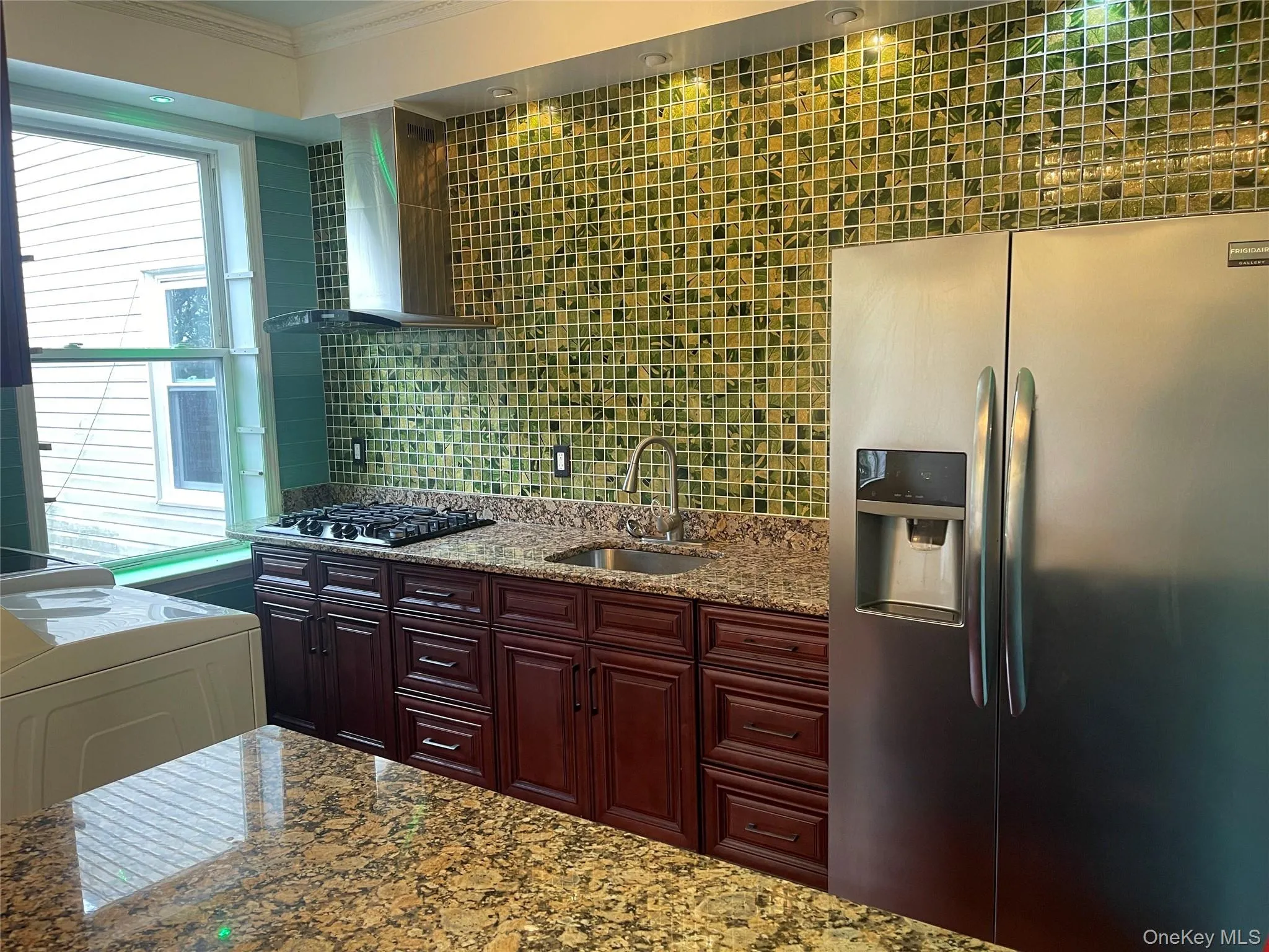 Kitchen featuring stainless steel fridge, tasteful backsplash, wall chimney exhaust hood, light stone countertops, and crown molding Kitchen featuring stainless steel fridge, tasteful backsplash, wall chimney exhaust hood, light stone countertops, and crown molding
